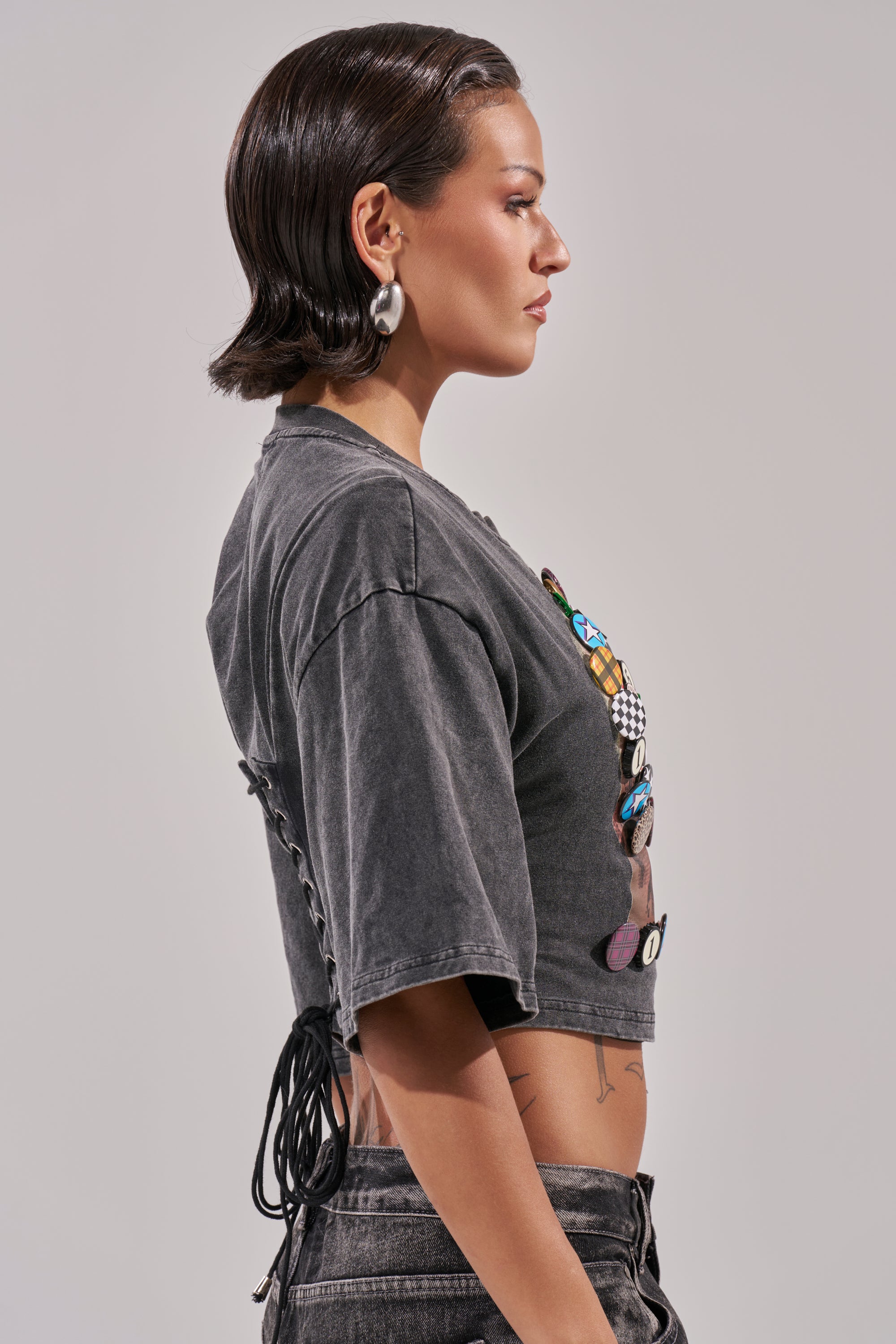 A woman with short, slicked-back dark hair is shown in profile wearing the PINNED DOWN Mineral Washed Tee in Grey, paired with black jeans. She accessorizes with large hoop earrings and displays visible arm and side tattoos.