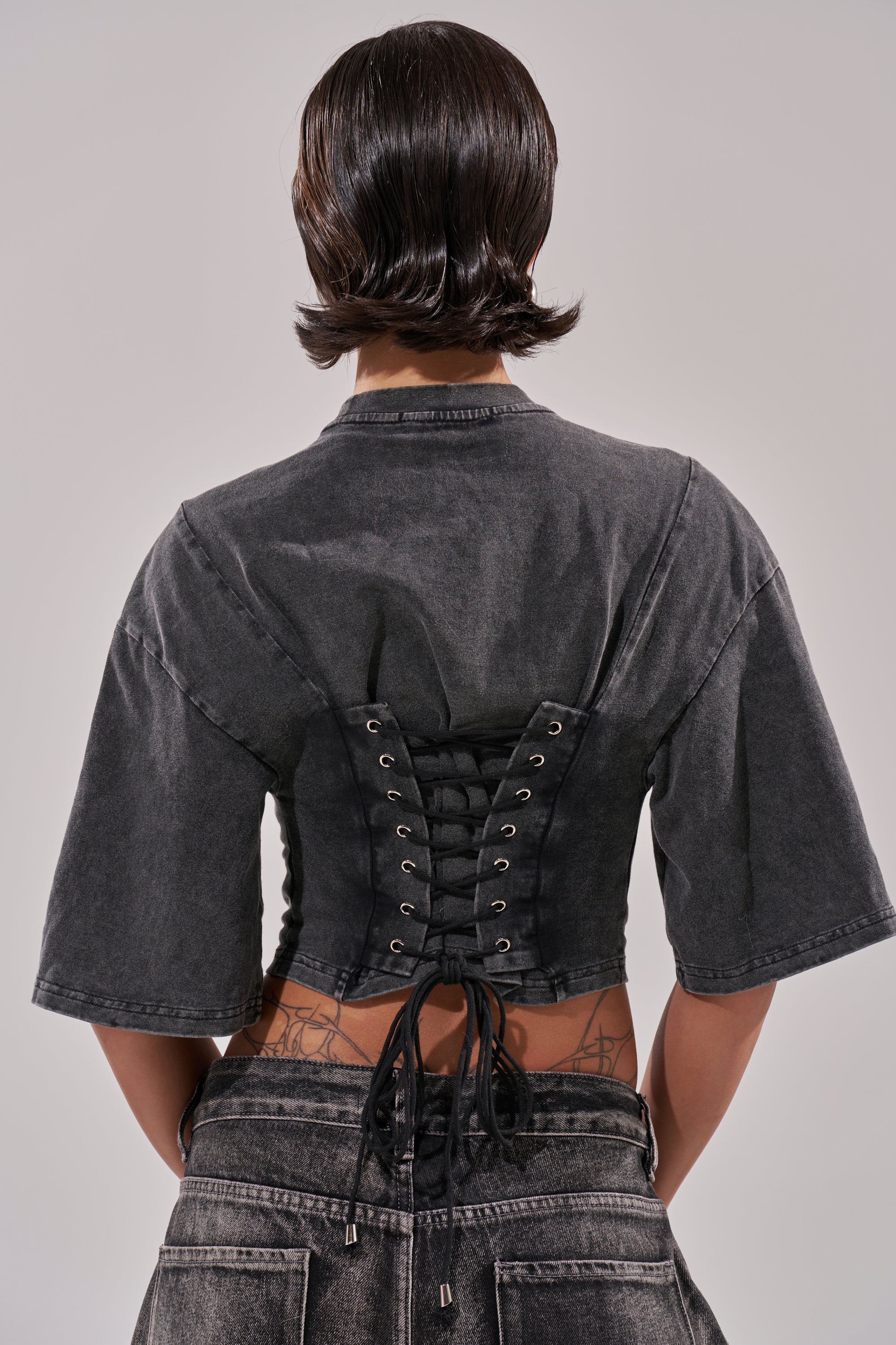 A person with short, dark, wavy hair is seen from behind wearing the PINNED DOWN MINERAL WASHED TEE IN GREY over high-waisted jeans. Some tattoos are visible on their lower back.