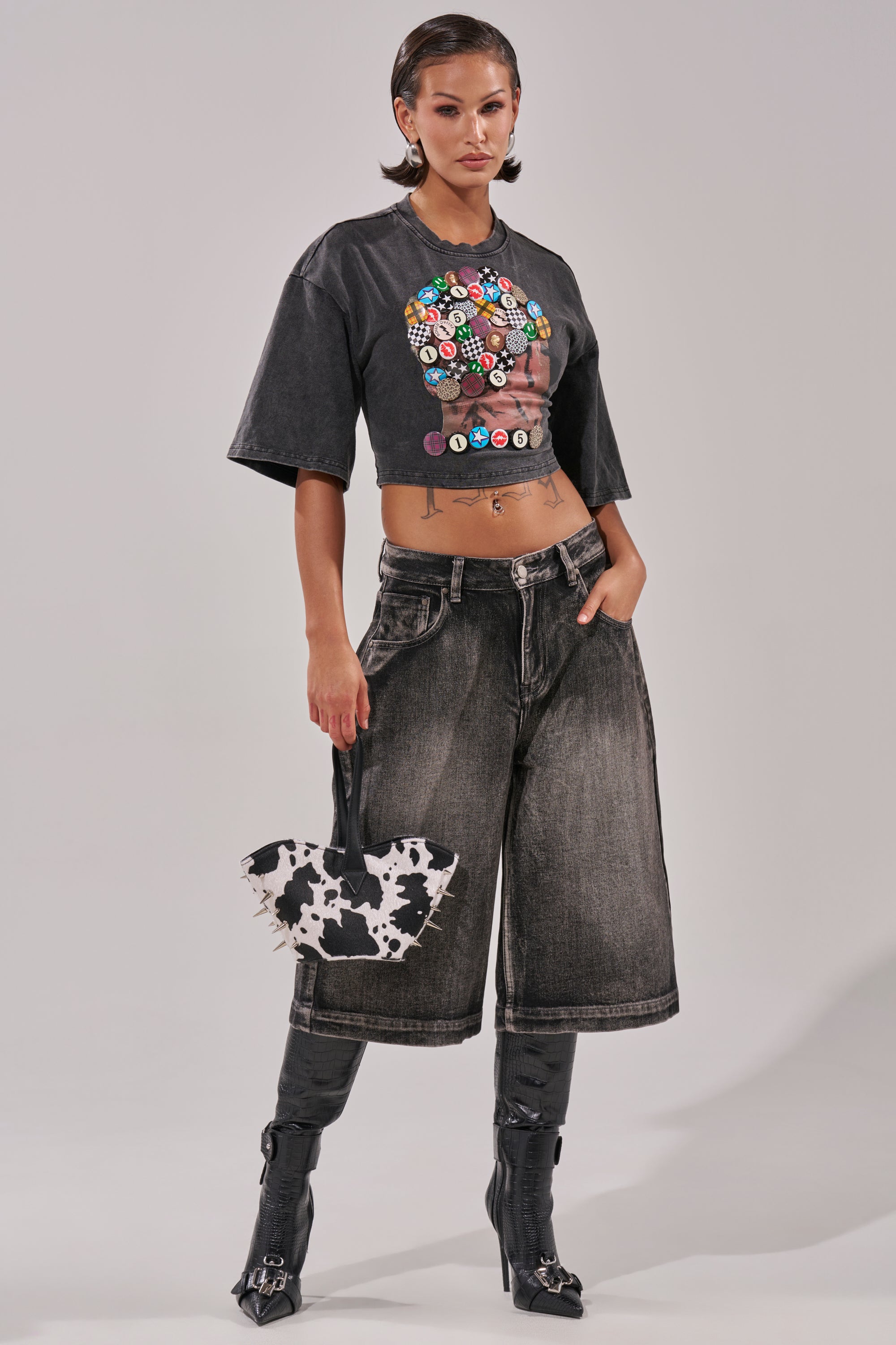 A woman poses in the PINNED DOWN MINERAL WASHED TEE IN GREY, wide-leg faded black jeans, black high-heeled boots, and a cow-print handbag. Her hair is slicked back as she stands against a plain background.