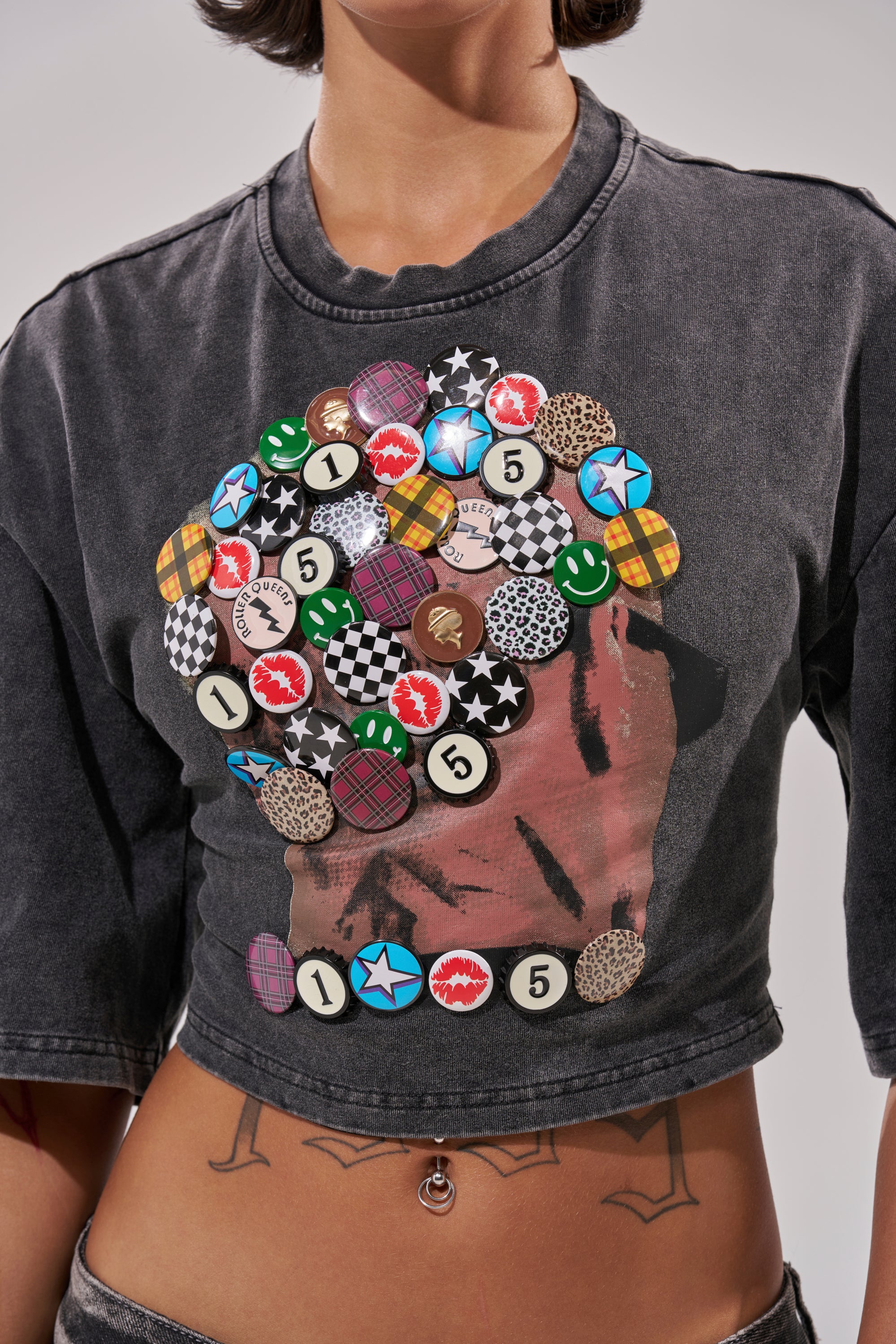 Wearing the PINNED DOWN MINERAL WASHED TEE IN GREY—a cropped, faded black top adorned with colorful pins and badges—they reveal a lower abdomen tattoo reading "IMEM.