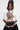 A woman with long dark hair wears the AKIRA Label PIN IT TO WIN IT TEE in a cropped silhouette, paired with a ruffled black skirt and a chunky, charm-adorned belt. She stands confidently against a plain light background.