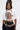A woman with long dark hair wears the AKIRA Label PIN IT TO WIN IT TEE, a cropped graphic top featuring a colorful pin-covered dog, styled with a beaded belt and black ruffled skirt against a plain background.