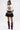 A woman stands against a plain backdrop wearing the PIN IT TO WIN IT TEE, a white cropped silhouette graphic top with colorful buttons, paired with a black ruffled mini skirt, multicolored charm belt, and black knee-high boots featuring red bows.