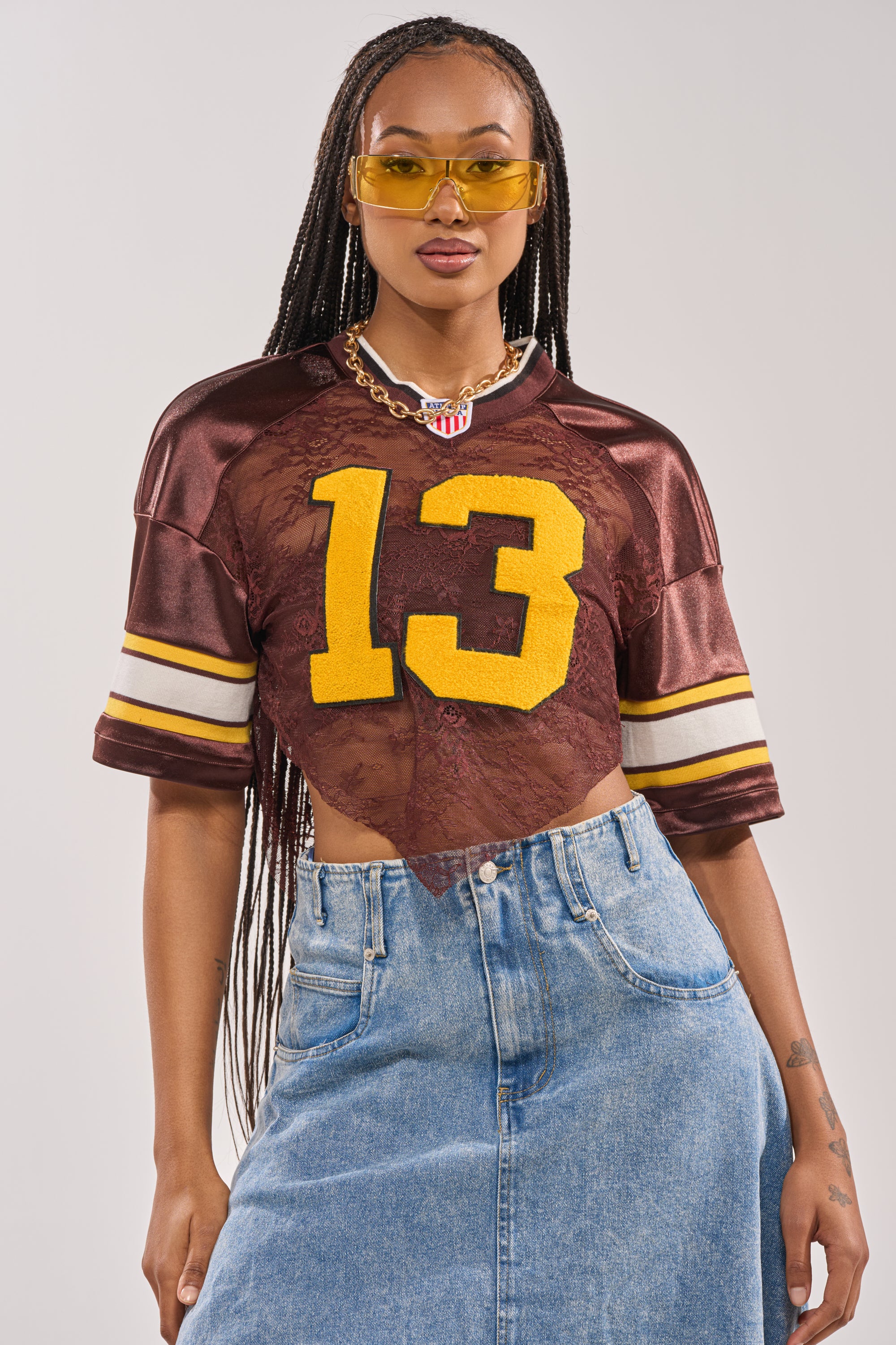 A woman wearing yellow sunglasses, a chain necklace, the LUCKY LACE JERSEY TOP IN BROWN with yellow number 13 over a sheer lace panel, and a denim skirt poses confidently against a plain background. 3