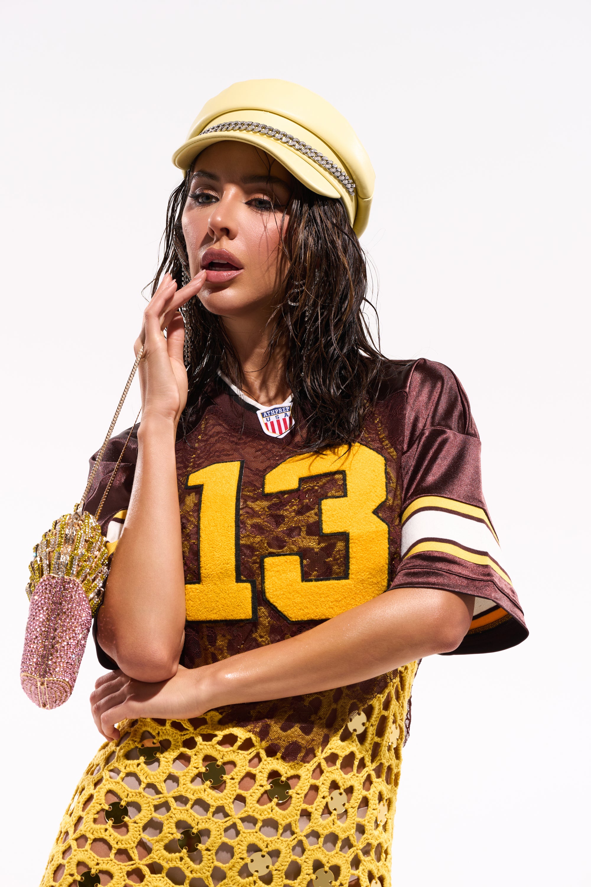 A woman with wet hair wears a varsity-style yellow hat, the LUCKY LACE JERSEY TOP IN BROWN with number 13, and a yellow crochet skirt. She holds a pink glittery ice cream cone-shaped bag, posing with her hand near her mouth. 0