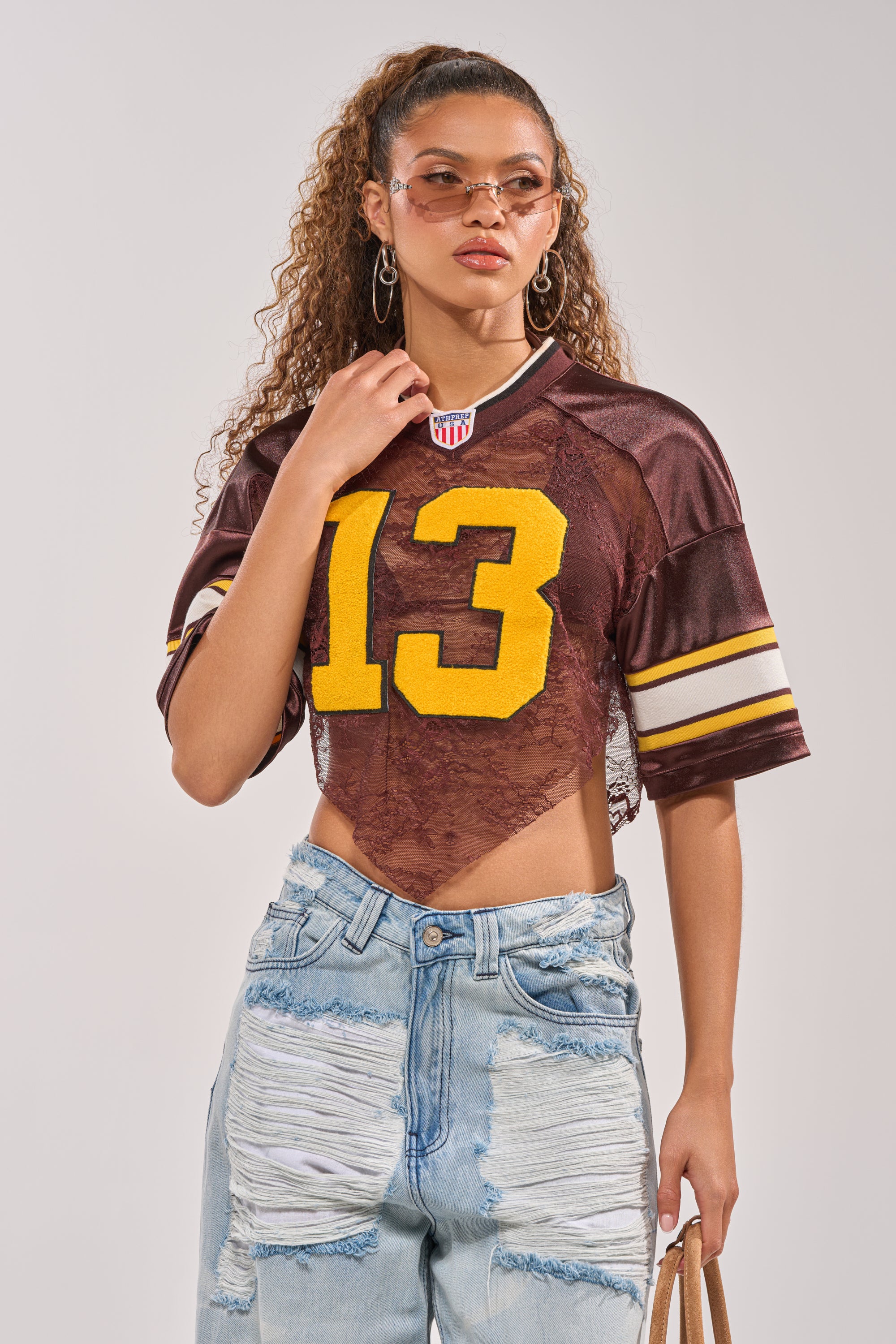 A woman wears the LUCKY LACE JERSEY TOP IN BROWN with varsity patch number 13, sheer lace fabric, paired with distressed light-wash jeans. She accessorizes with hoop earrings, sunglasses, curly hair, and a brown tote bag. 0