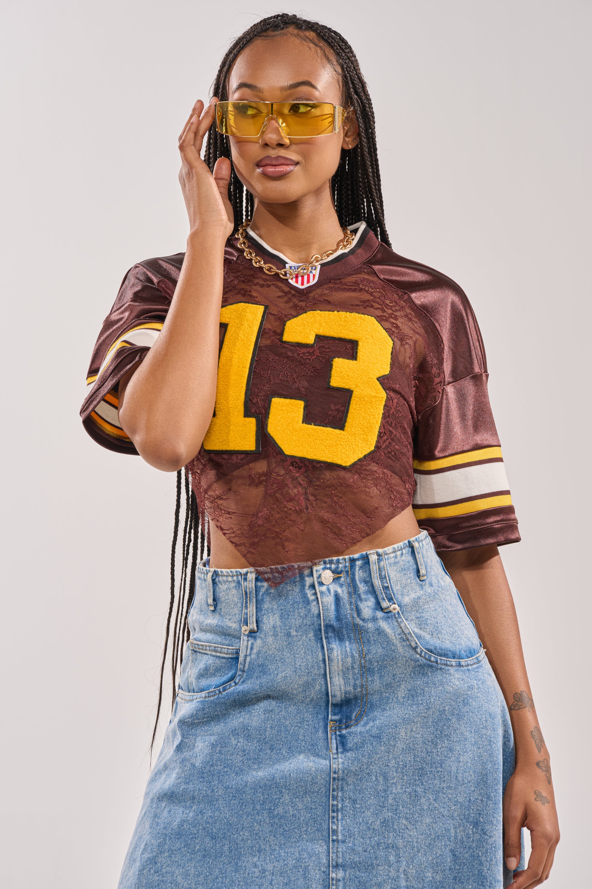 A woman wearing yellow sunglasses, a necklace, a denim skirt, and the LUCKY LACE JERSEY TOP IN BROWN poses confidently against a light background. 4
