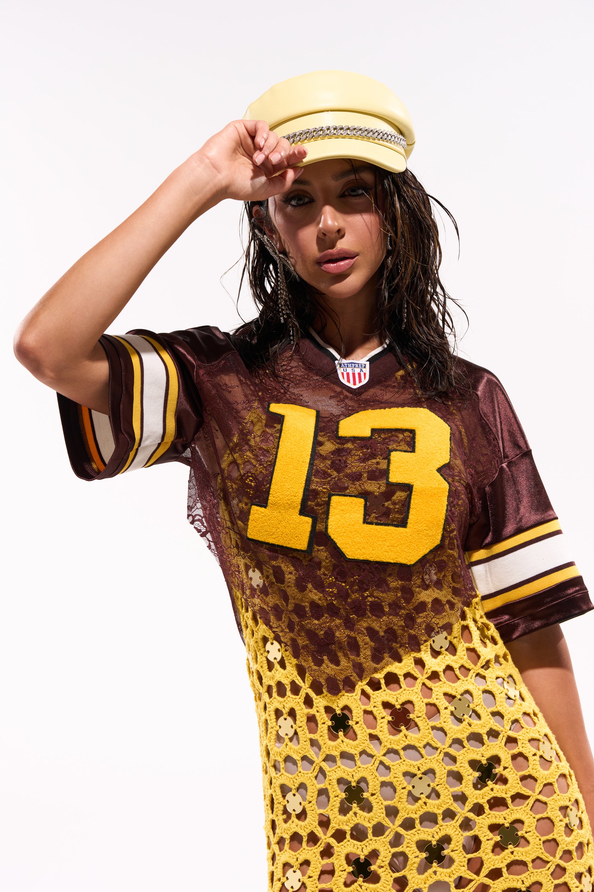 A woman in a yellow cap wears the LUCKY LACE JERSEY TOP IN BROWN, featuring the number 13 and transitioning into a yellow, sheer floral lace skirt at the bottom, standing against a white background. 1