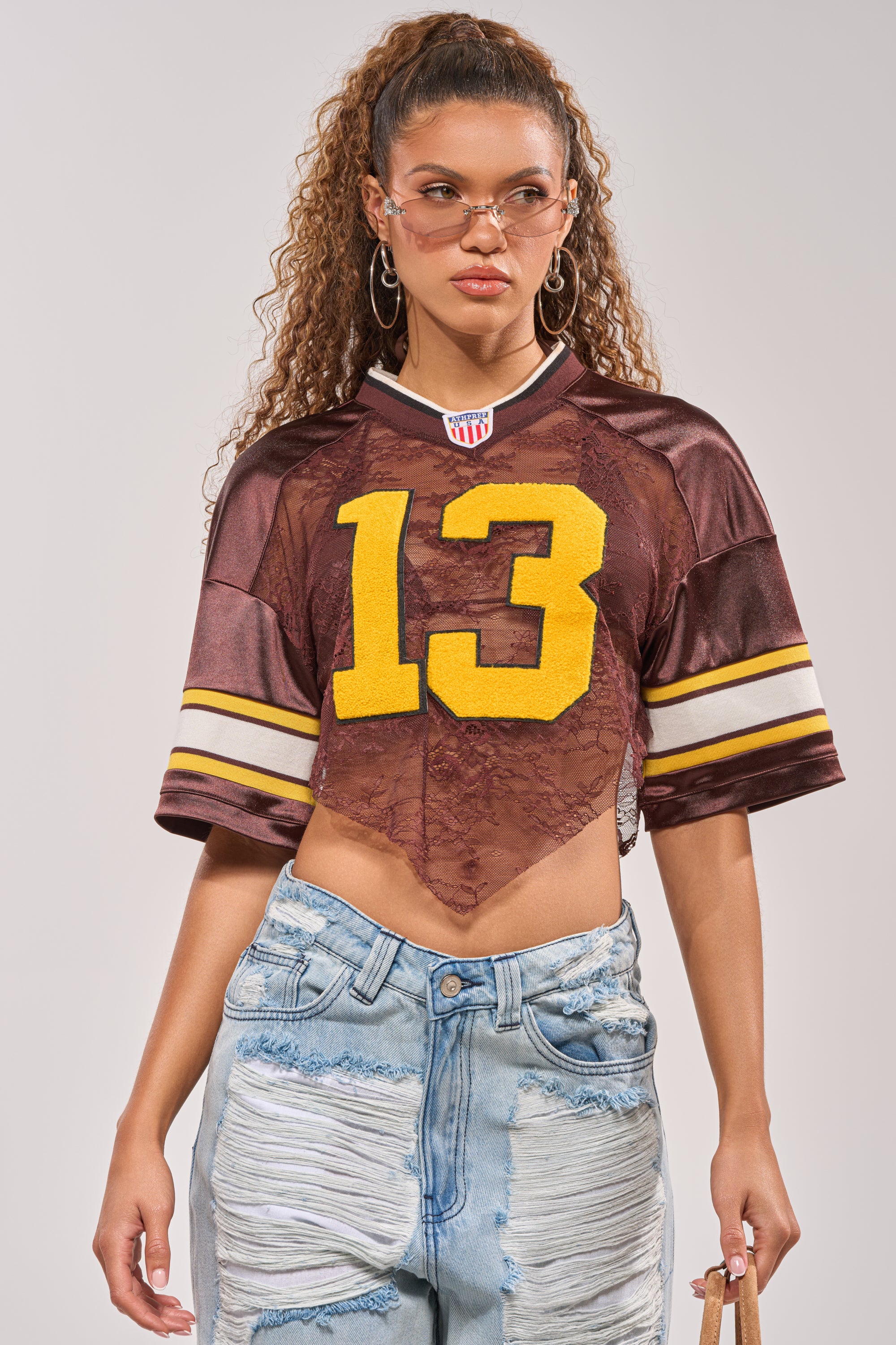 A woman in the LUCKY LACE JERSEY TOP IN BROWN with yellow "13", ripped light blue jeans, hoop earrings, and tinted glasses poses against a plain background. 1