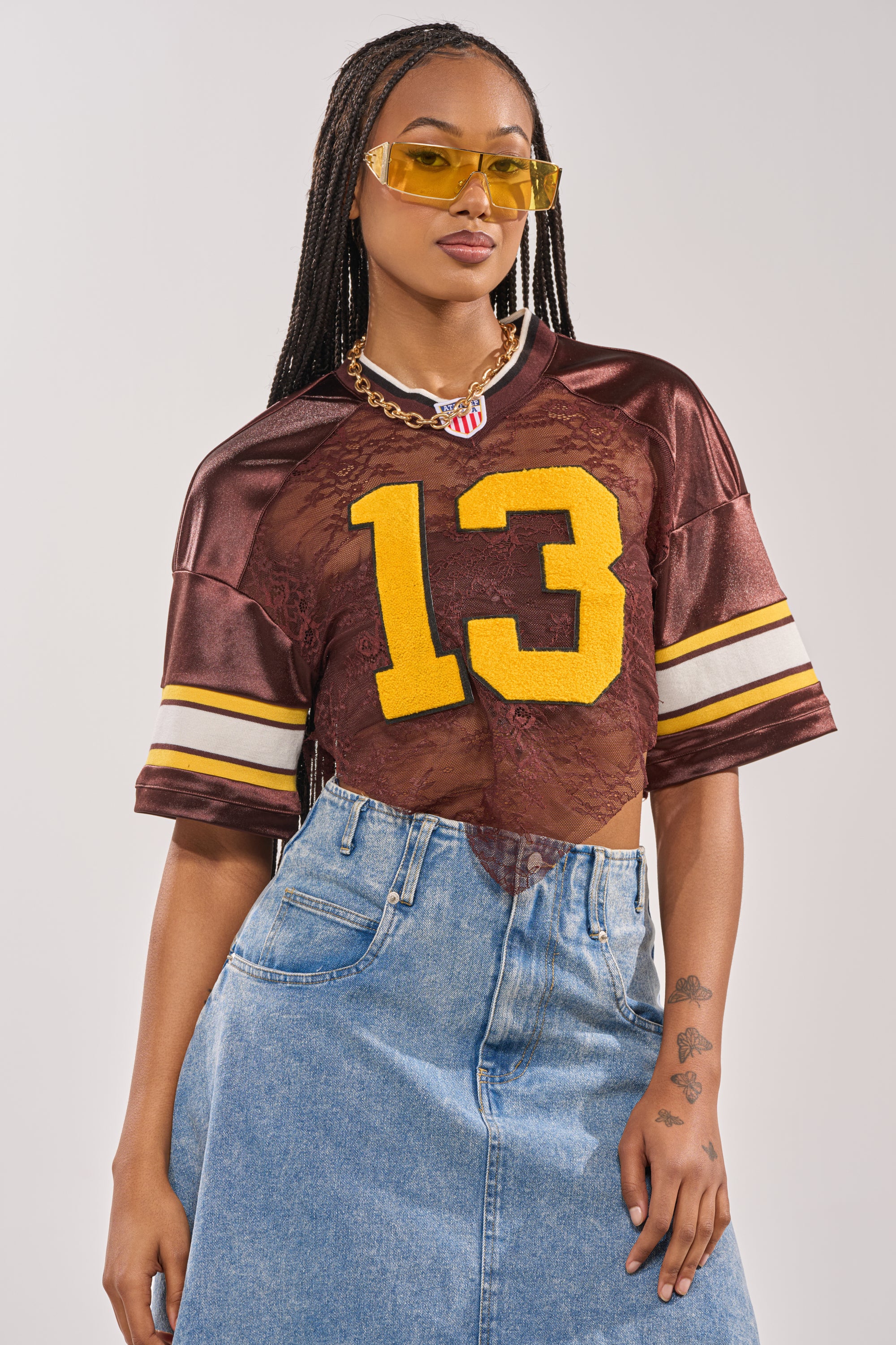 A woman poses confidently in the LUCKY LACE JERSEY TOP IN BROWN, featuring a varsity style with a bold yellow number 13 over floral lace, paired with yellow sunglasses, a chunky necklace, and high-waisted blue denim jeans. 5