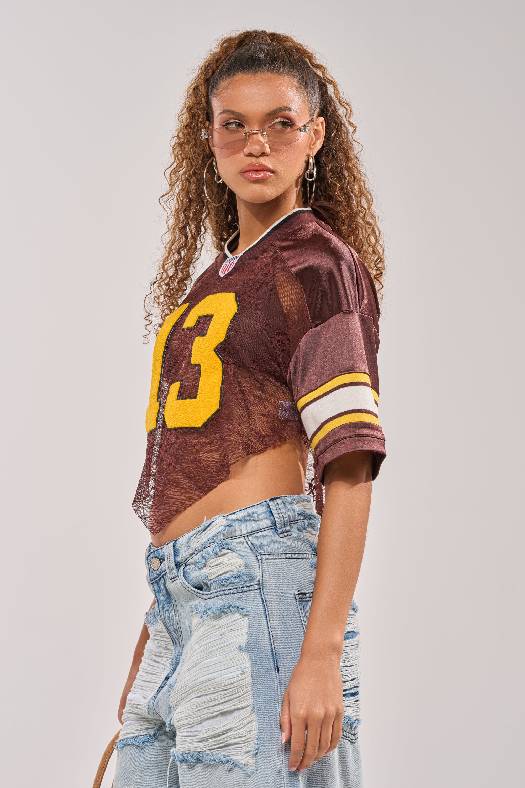 A woman with curly hair wears small sunglasses, hoop earrings, the LUCKY LACE JERSEY TOP IN BROWN featuring a yellow "13," and light blue ripped jeans, standing with one hand at her side against a plain background. 2
