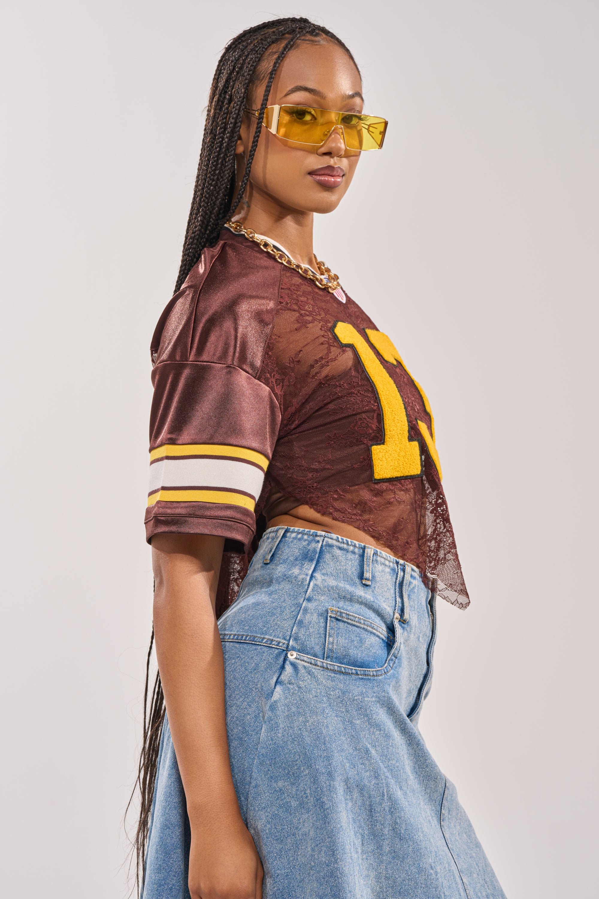 A woman with long braids wears yellow-tinted sunglasses, a gold chain, high-waisted blue jeans, and the LUCKY LACE JERSEY TOP IN BROWN featuring varsity patch details. She stands in profile against a neutral background. 6