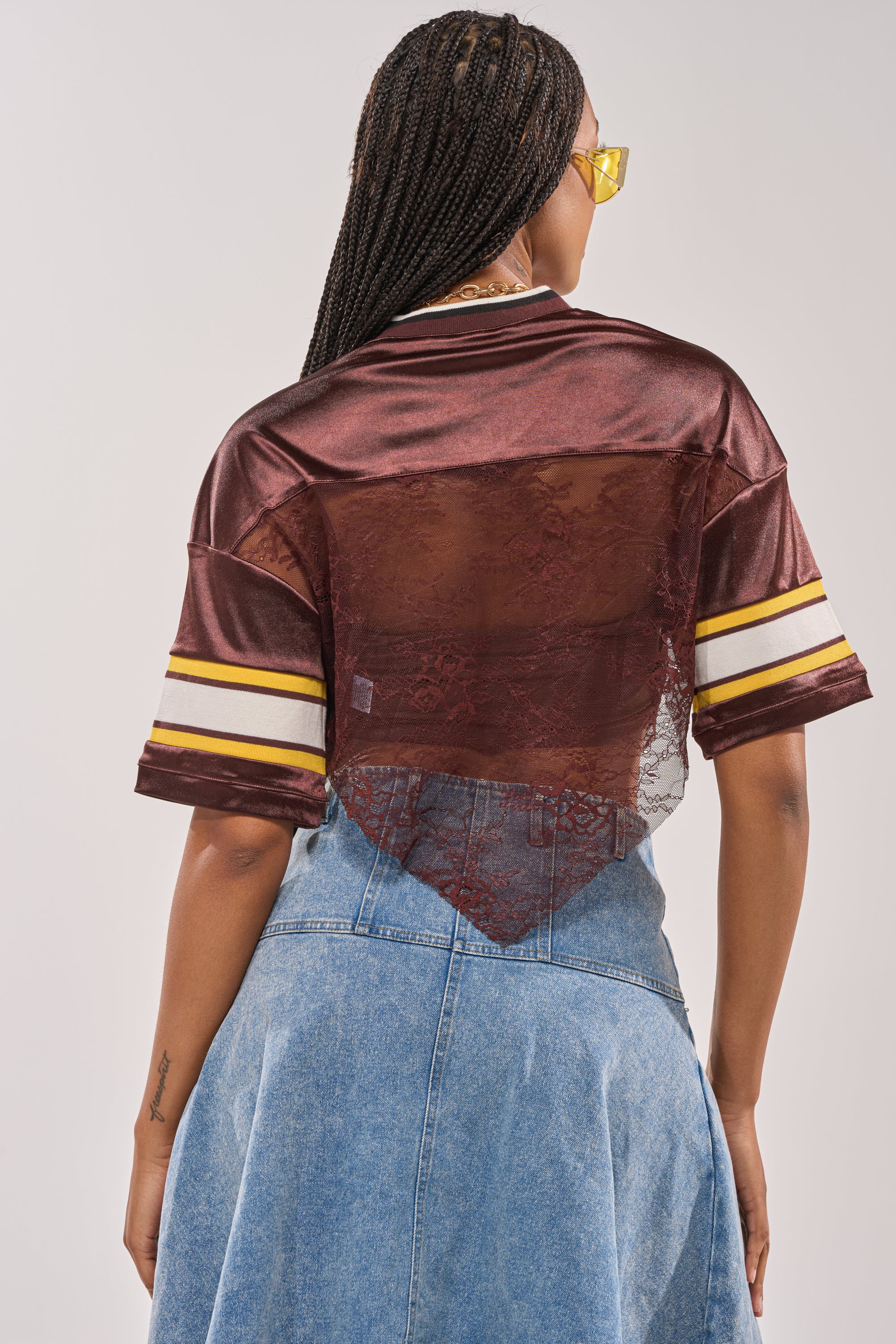 A woman with braided hair, wearing yellow-tinted glasses, faces away from the camera in the LUCKY LACE JERSEY TOP IN BROWN, which features a shiny finish, sheer floral lace back panel, striped short sleeves, and a high-waisted denim skirt. 7