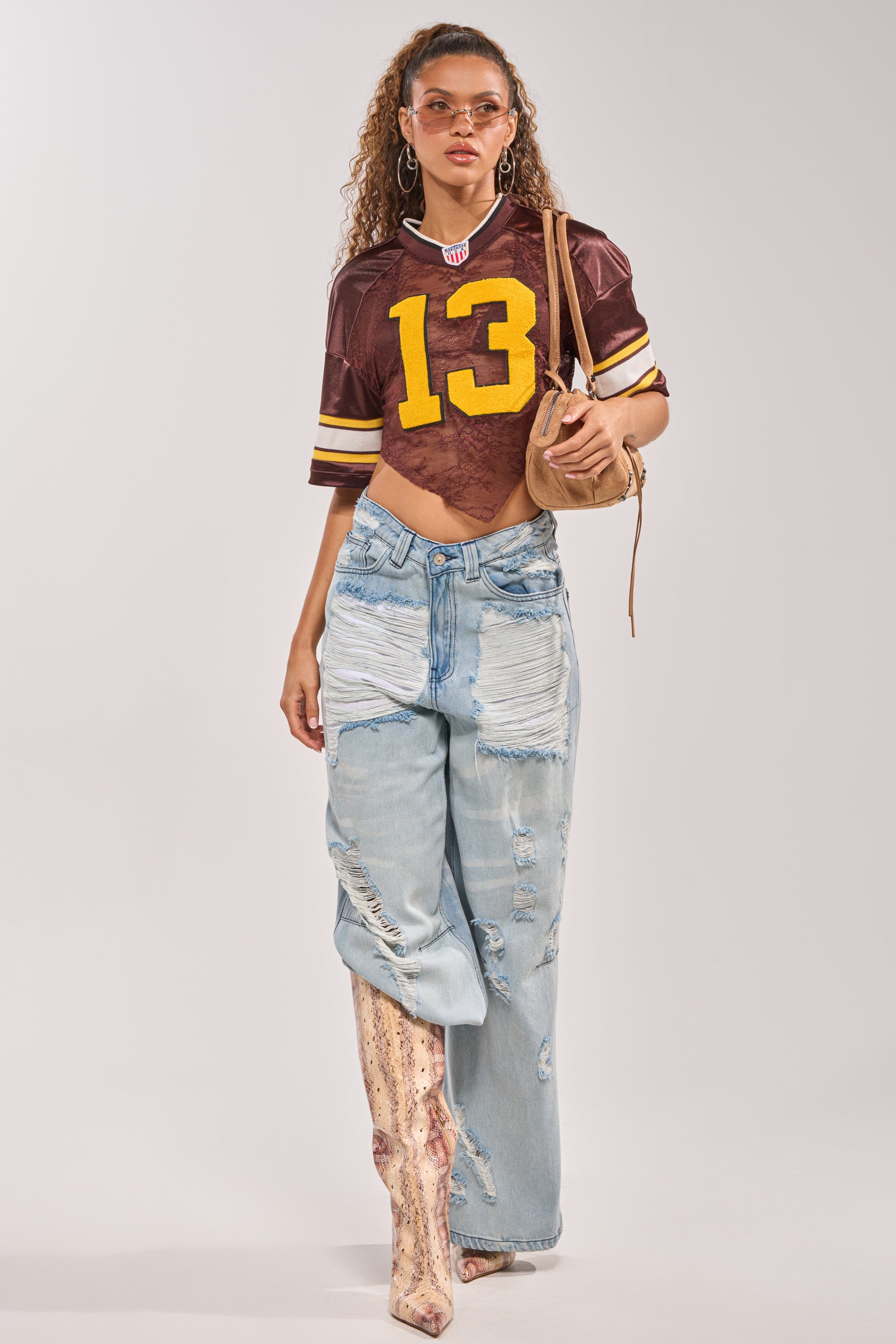 A woman sports tinted sunglasses, the LUCKY LACE JERSEY TOP IN BROWN with the number 13, ripped light-wash patchwork jeans, knee-high snakeskin boots, hoop earrings, and a tan shoulder bag. 4