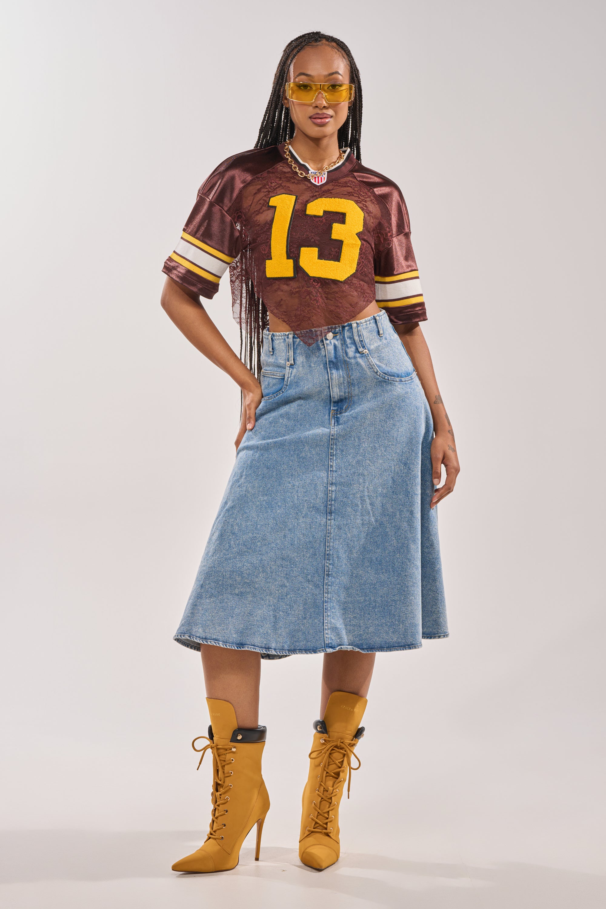 A woman poses confidently in the LUCKY LACE JERSEY TOP IN BROWN with the number 13 and varsity patch, paired with a blue denim midi skirt, mustard lace-up heeled boots, yellow sunglasses, and a necklace against a plain background. 8