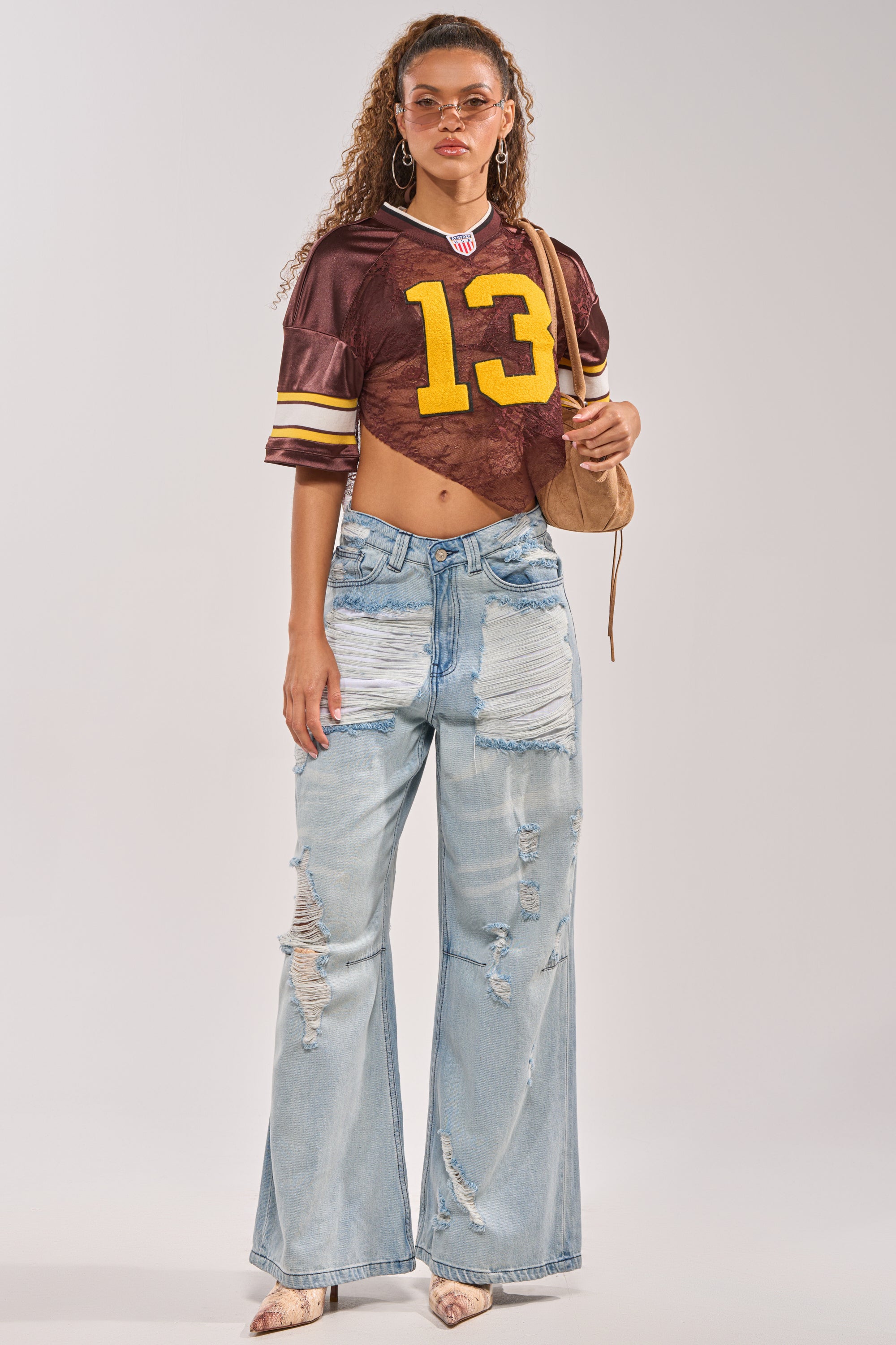 A woman in the LUCKY LACE JERSEY TOP IN BROWN, light blue distressed wide-leg jeans, pointed shoes, hoop earrings, sunglasses, and a small tan shoulder bag stands against a plain background. 5