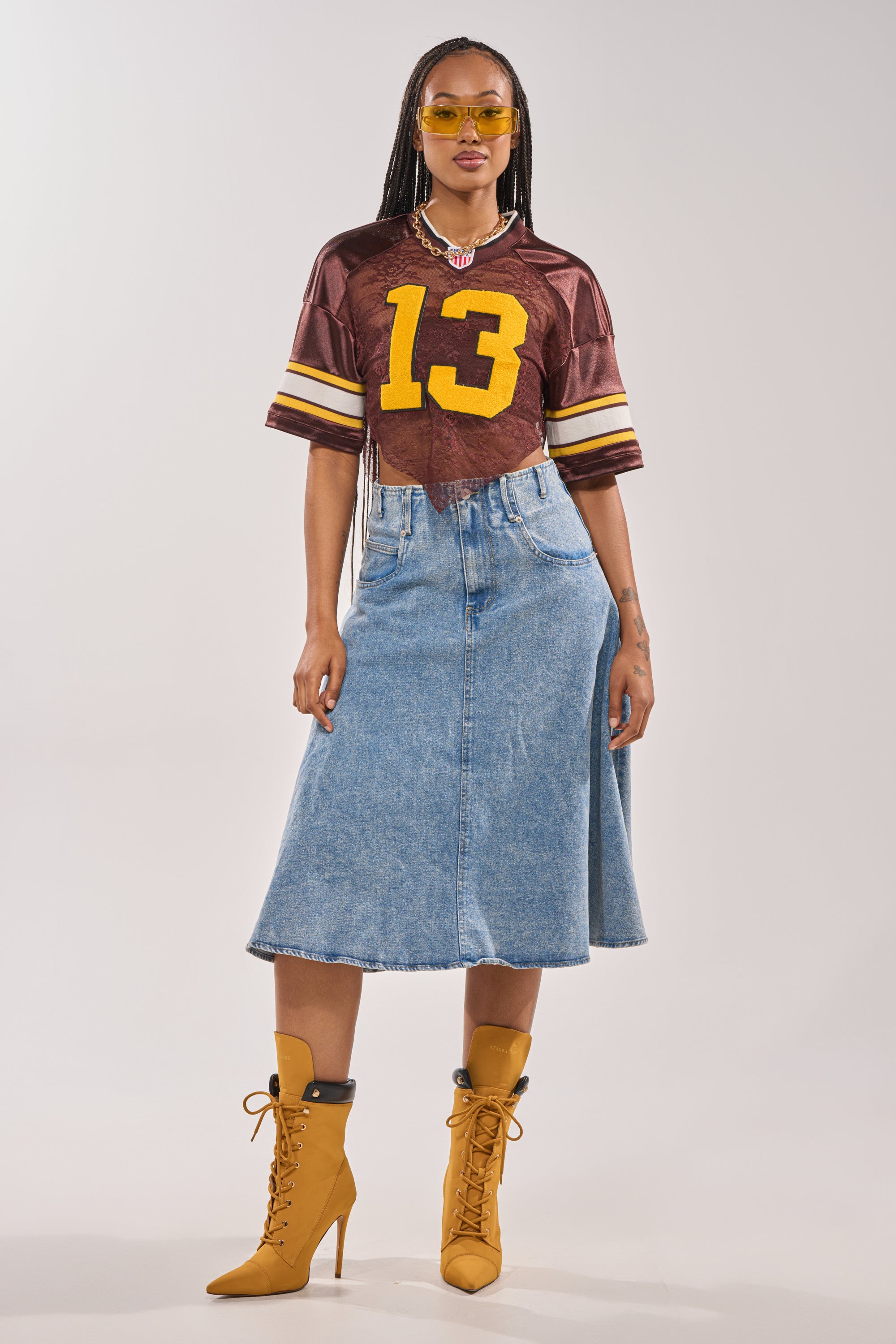 A woman in yellow sunglasses, the LUCKY LACE JERSEY TOP IN BROWN with “13”, a knee-length denim skirt, and mustard lace-up heeled boots stands confidently against a plain background. 9