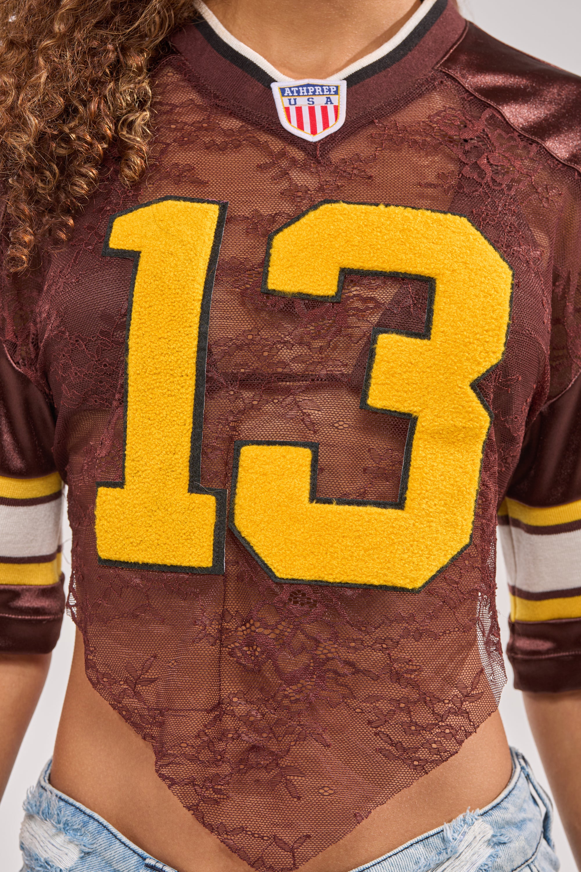 A person wears the LUCKY LACE JERSEY TOP IN BROWN featuring a floral lace panel and yellow number 13, paired with light denim shorts. The image is cropped at the shoulders and waist. 9