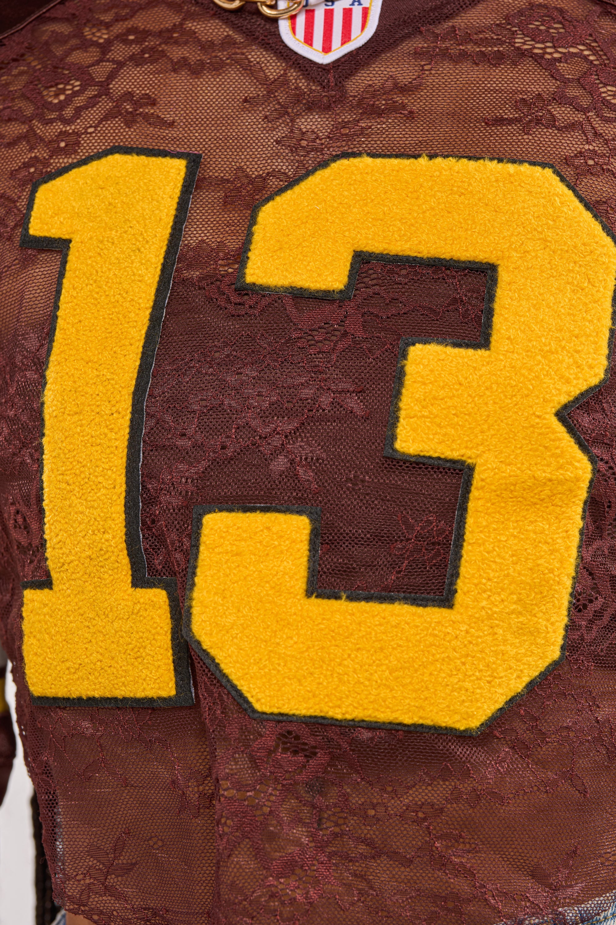 A close-up of the LUCKY LACE JERSEY TOP IN BROWN shows its brown floral lace panel with large yellow and black varsity-style "13" numbers and a small American flag-inspired patch at the top. 10
