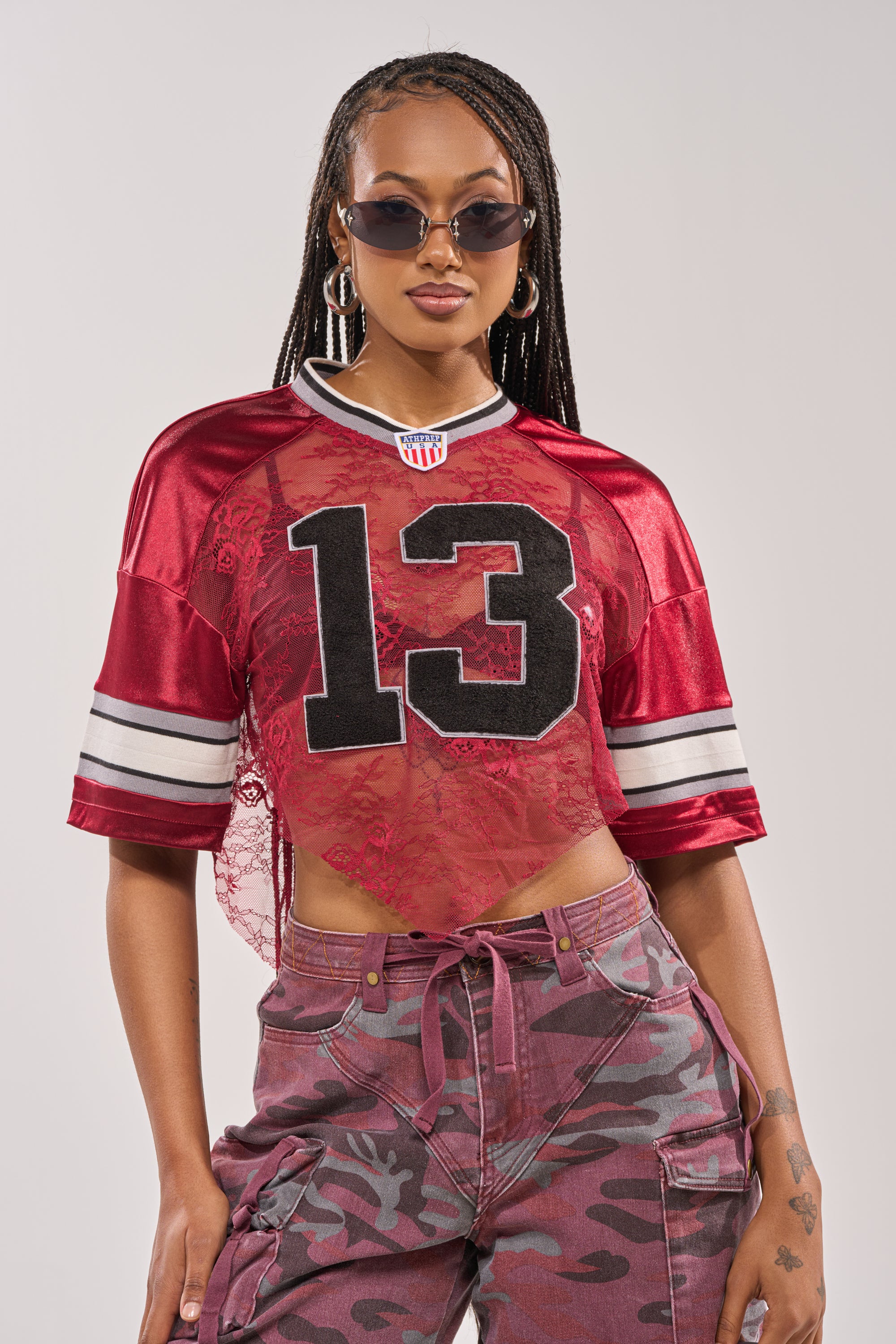 A woman in sunglasses, hoop earrings, camo cargo pants, and the LUCKY LACE JERSEY TOP IN BURGUNDY with a number 13 design poses confidently against a plain background. 2