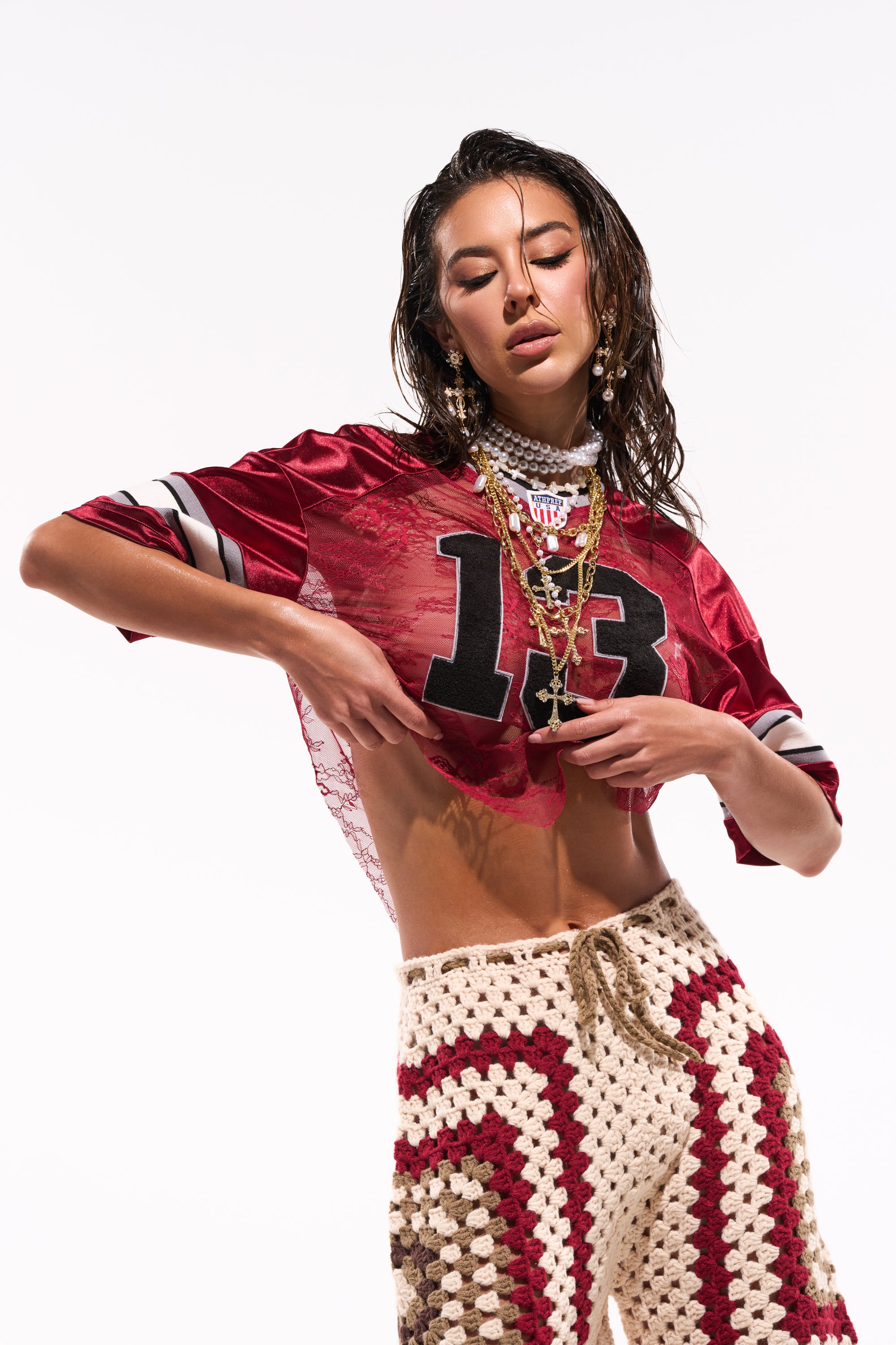 A woman poses in the LUCKY LACE JERSEY TOP IN BURGUNDY, featuring sheer red lace with the number 13. She pairs it with high-waisted crocheted pants, layered necklaces, and wet-styled hair against a white background. 1