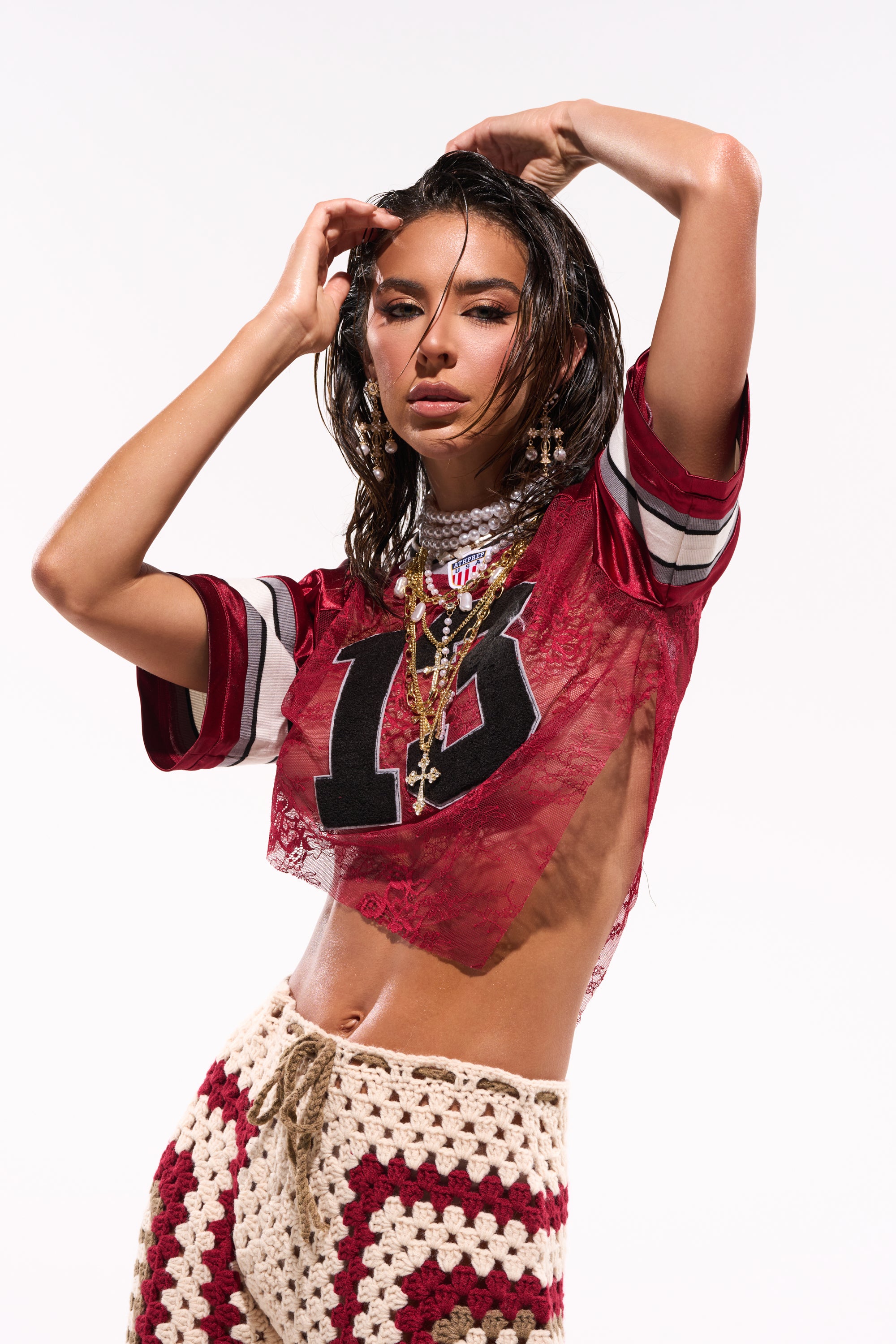 A woman poses confidently against a white background in the LUCKY LACE JERSEY TOP IN BURGUNDY, styled with layered necklaces and crocheted pants. Her wet, tousled hair frames her as she holds her hands near her head. 0