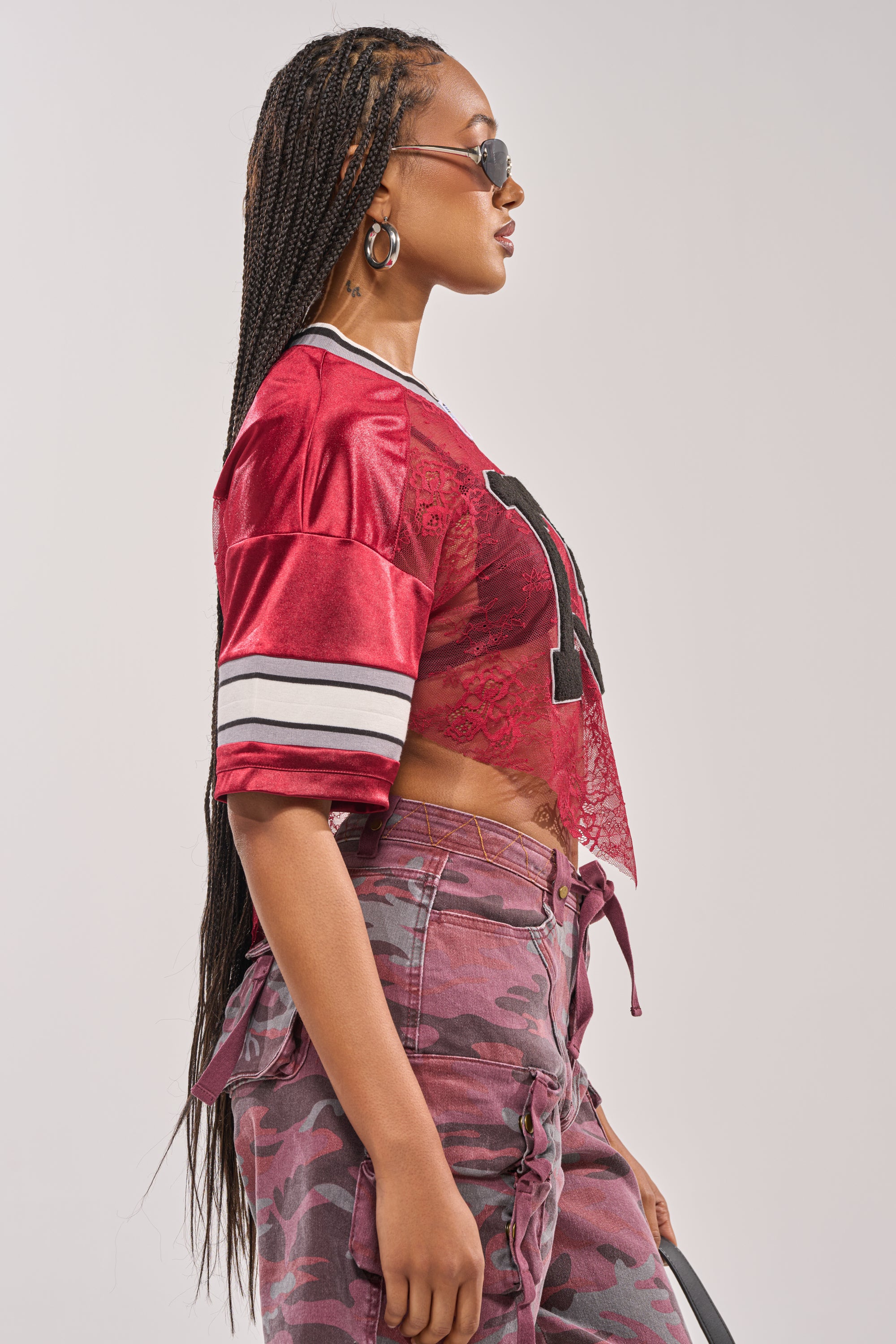 A woman with long braided hair stands in profile against a plain background, wearing sunglasses, hoop earrings, pink camo pants, and the LUCKY LACE JERSEY TOP IN BURGUNDY with sheer red lace details. 4