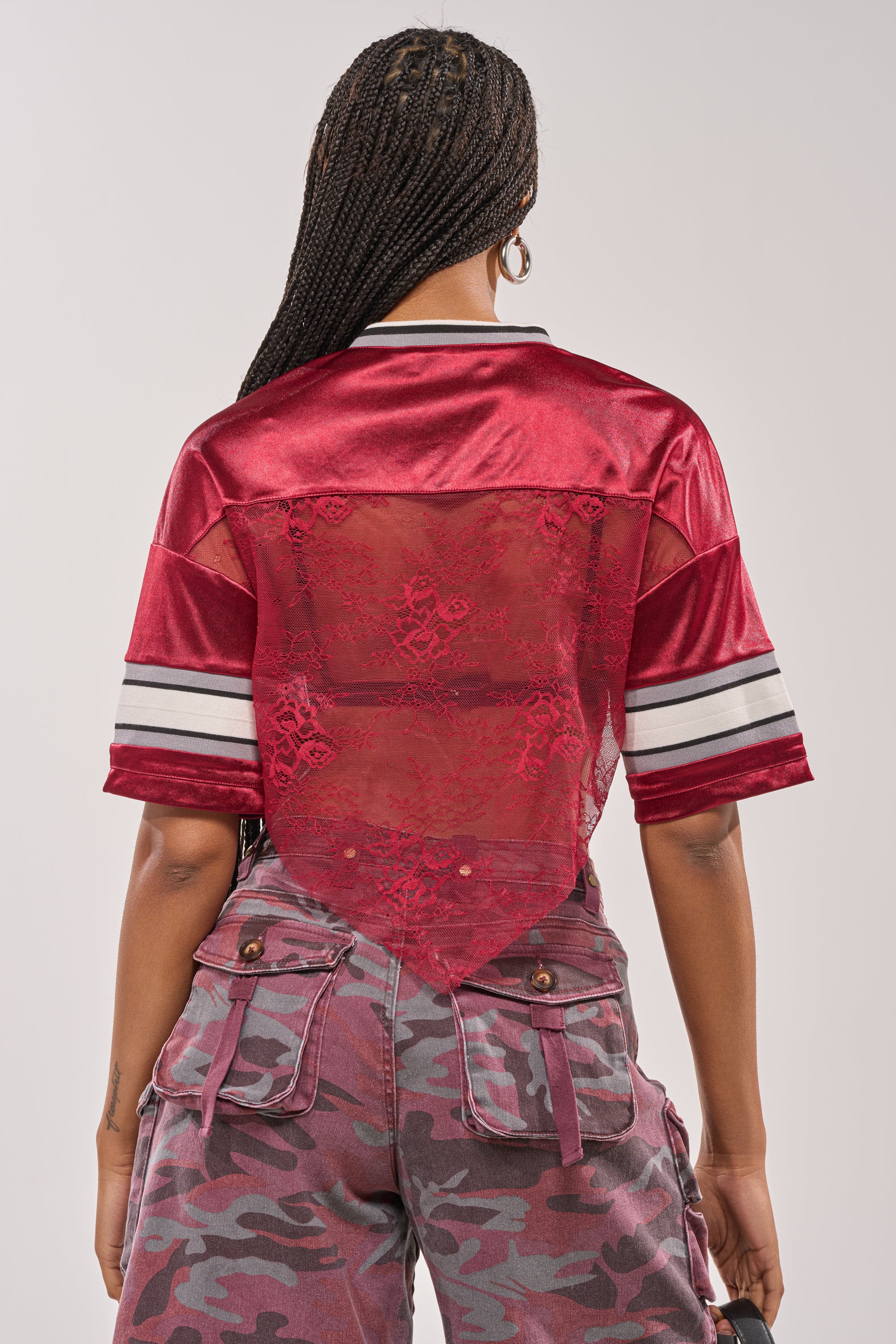 A person with long braids is seen from the back wearing the LUCKY LACE JERSEY TOP IN BURGUNDY with striped sleeves and purple camouflage cargo pants. 5