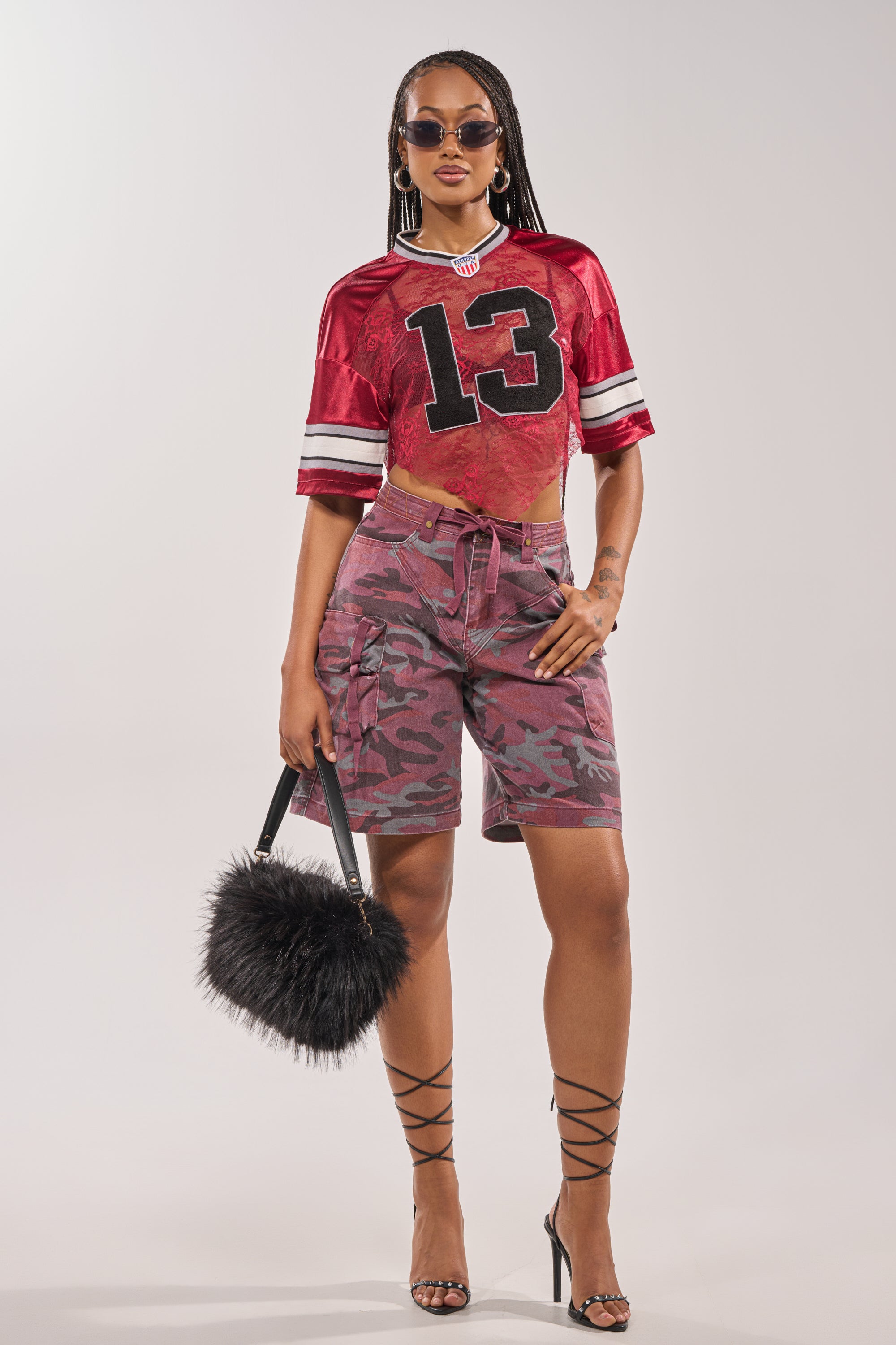 A woman in the LUCKY LACE JERSEY TOP IN BURGUNDY with camo shorts, strappy heels, sunglasses, hoop earrings, and a black furry handbag stands confidently against a plain background. 6
