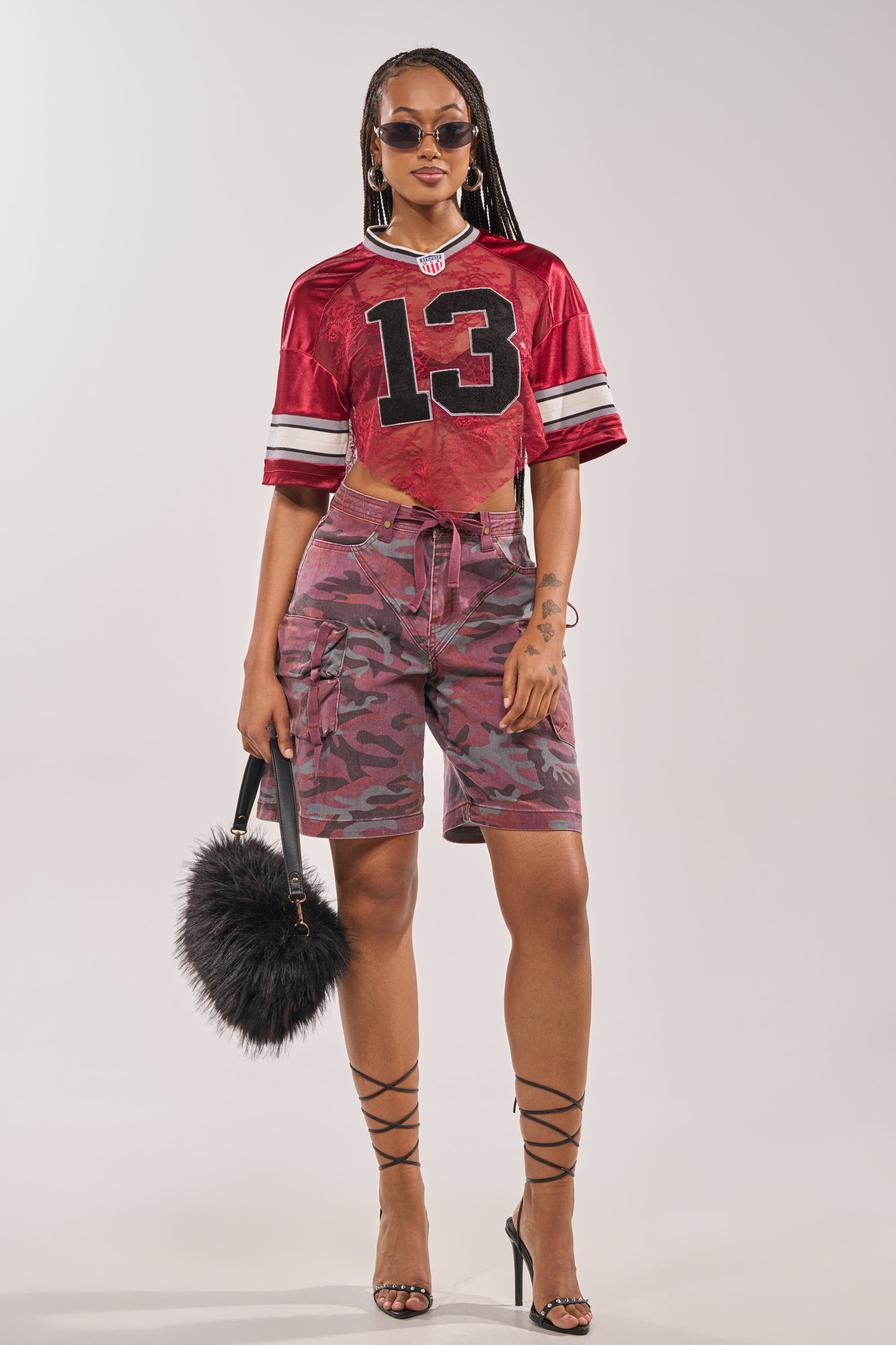 A woman stands confidently in the LUCKY LACE JERSEY TOP IN BURGUNDY, sunglasses, pink camo cargo shorts, lace-up high heels, and holding a black furry handbag. 7