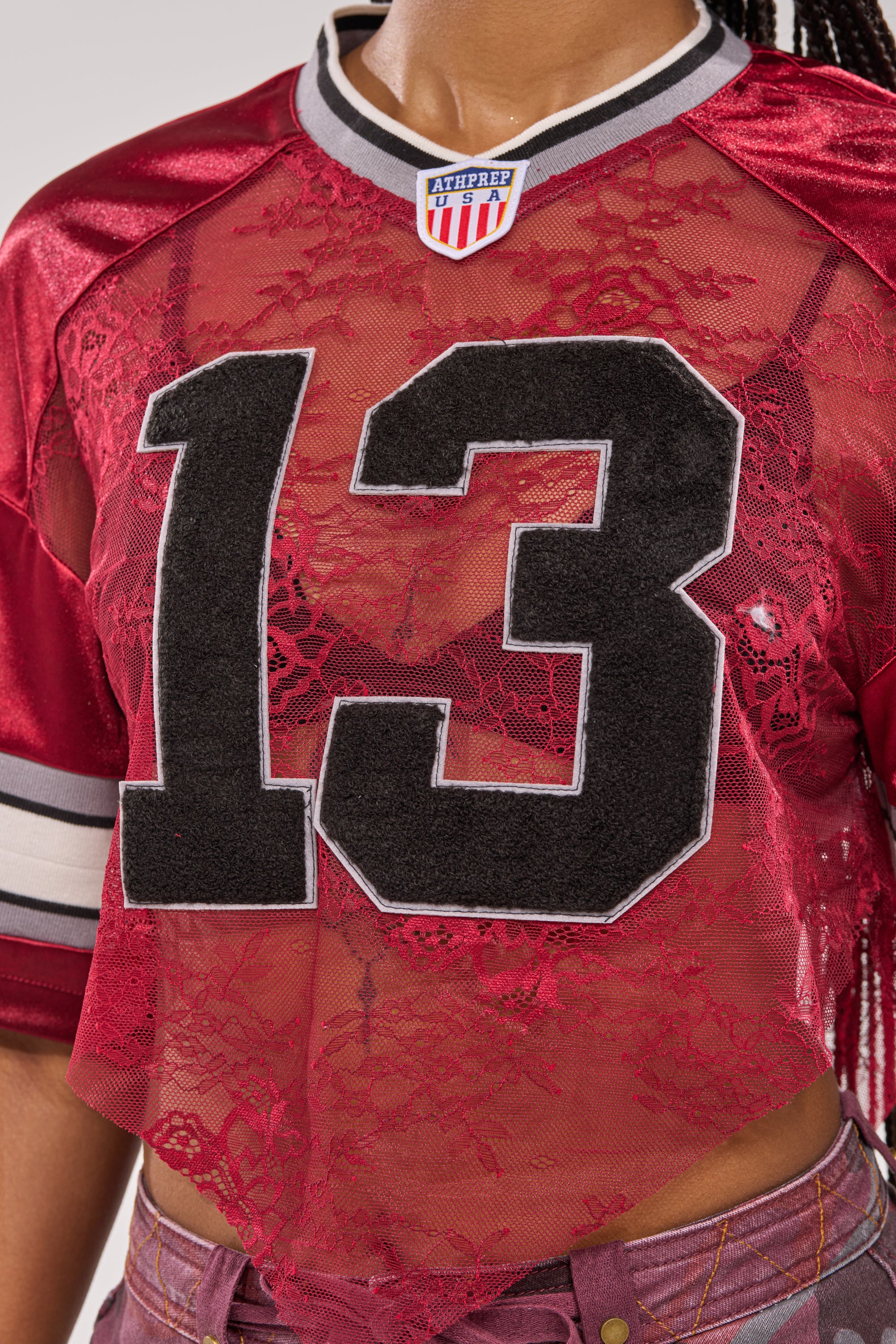 A person wears the LUCKY LACE JERSEY TOP IN BURGUNDY—a sheer red lace sports-style jersey with bold black and white 13, an athletic collar patch, and grey and white striped sleeves. 8