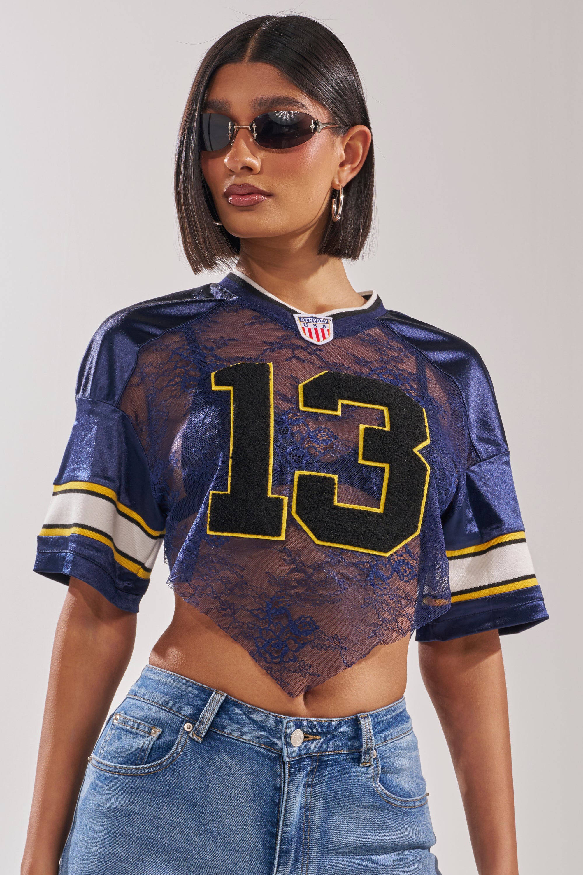 A woman with a short bob and sunglasses models the LUCKY LACE JERSEY TOP IN NAVY, featuring a varsity cropped style with the number 13 and sheer lace details, paired with blue jeans against a plain light background. 0