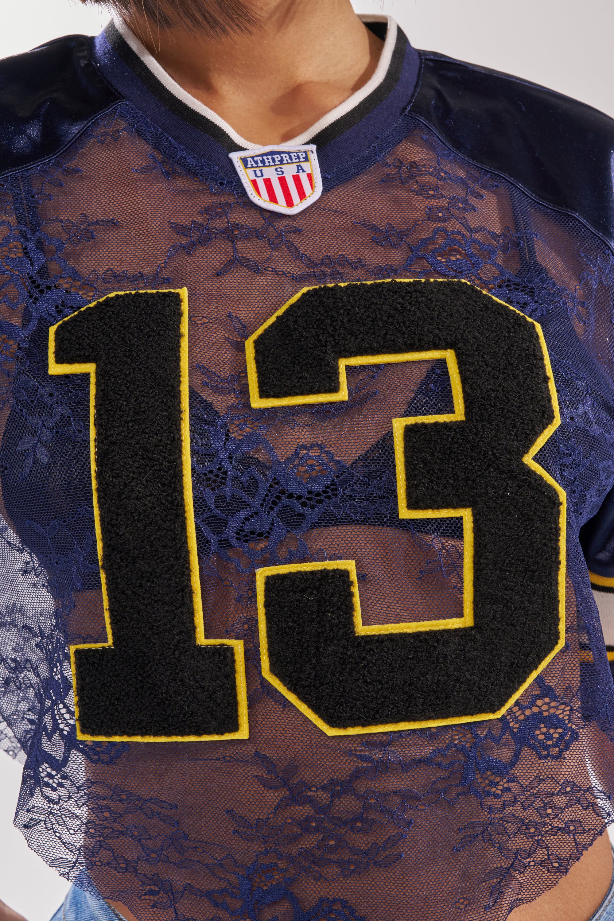 Someone is wearing the LUCKY LACE JERSEY TOP IN NAVY, a sheer lace cropped varsity-style shirt with a bold black and yellow number 13 and a small football-inspired shield patch near the collar. 9