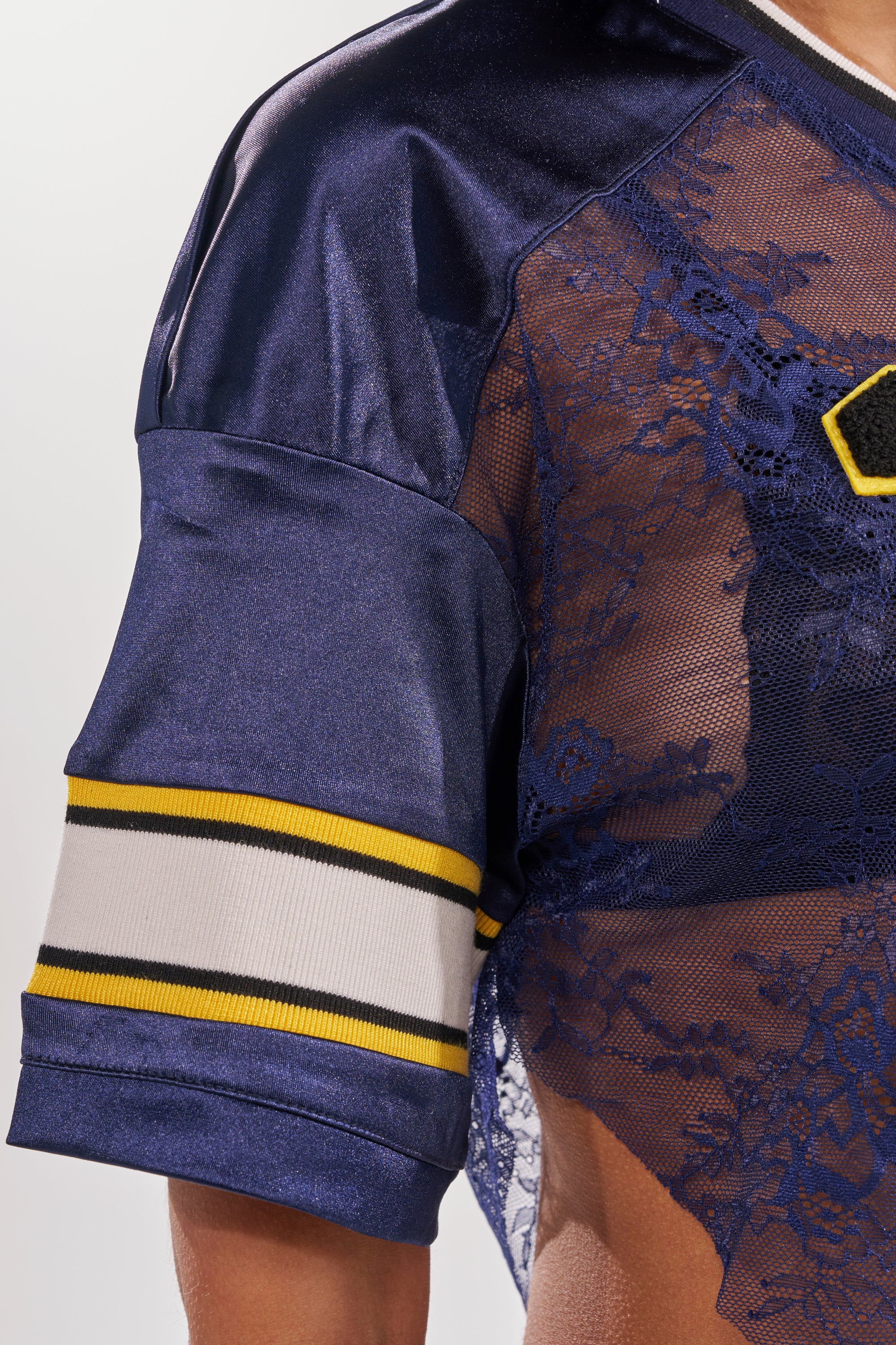A close-up of a person wearing the LUCKY LACE JERSEY TOP IN NAVY, featuring sheer navy lace and yellow and white striped sleeve trim. This cropped varsity top pairs sporty vibes with elegant style, revealing a hint of skin underneath. 10