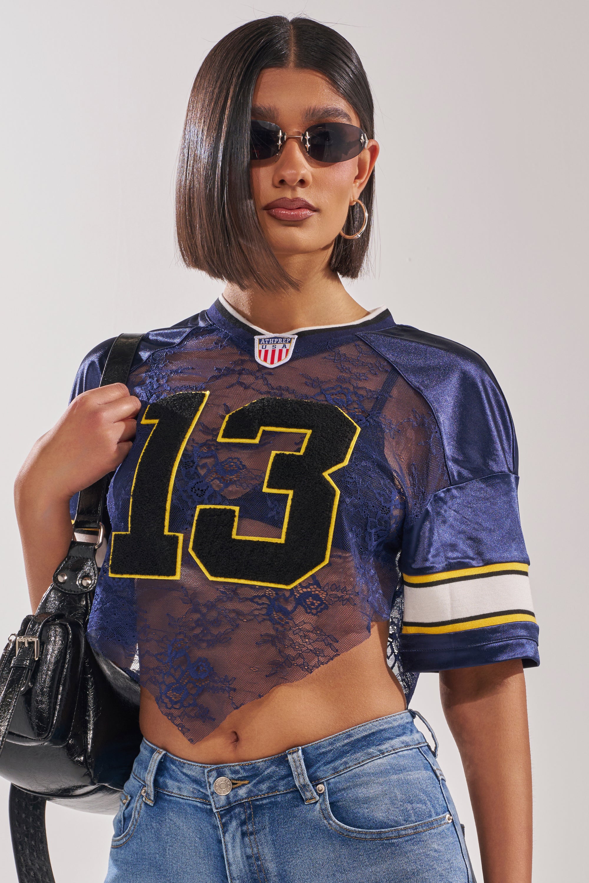 A woman with straight dark hair wears sunglasses, hoop earrings, blue jeans, a black shoulder bag, and the LUCKY LACE JERSEY TOP IN NAVY featuring sheer lace and a bold yellow "13" with football stripes. 1