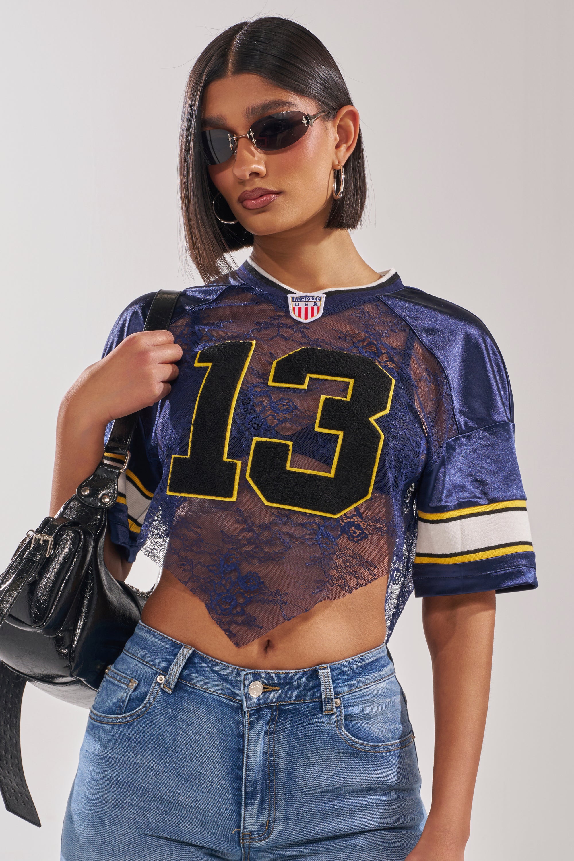 A woman in sunglasses and blue jeans poses confidently against a plain background, wearing the LUCKY LACE JERSEY TOP IN NAVY with the number 13 and carrying a black handbag. 2