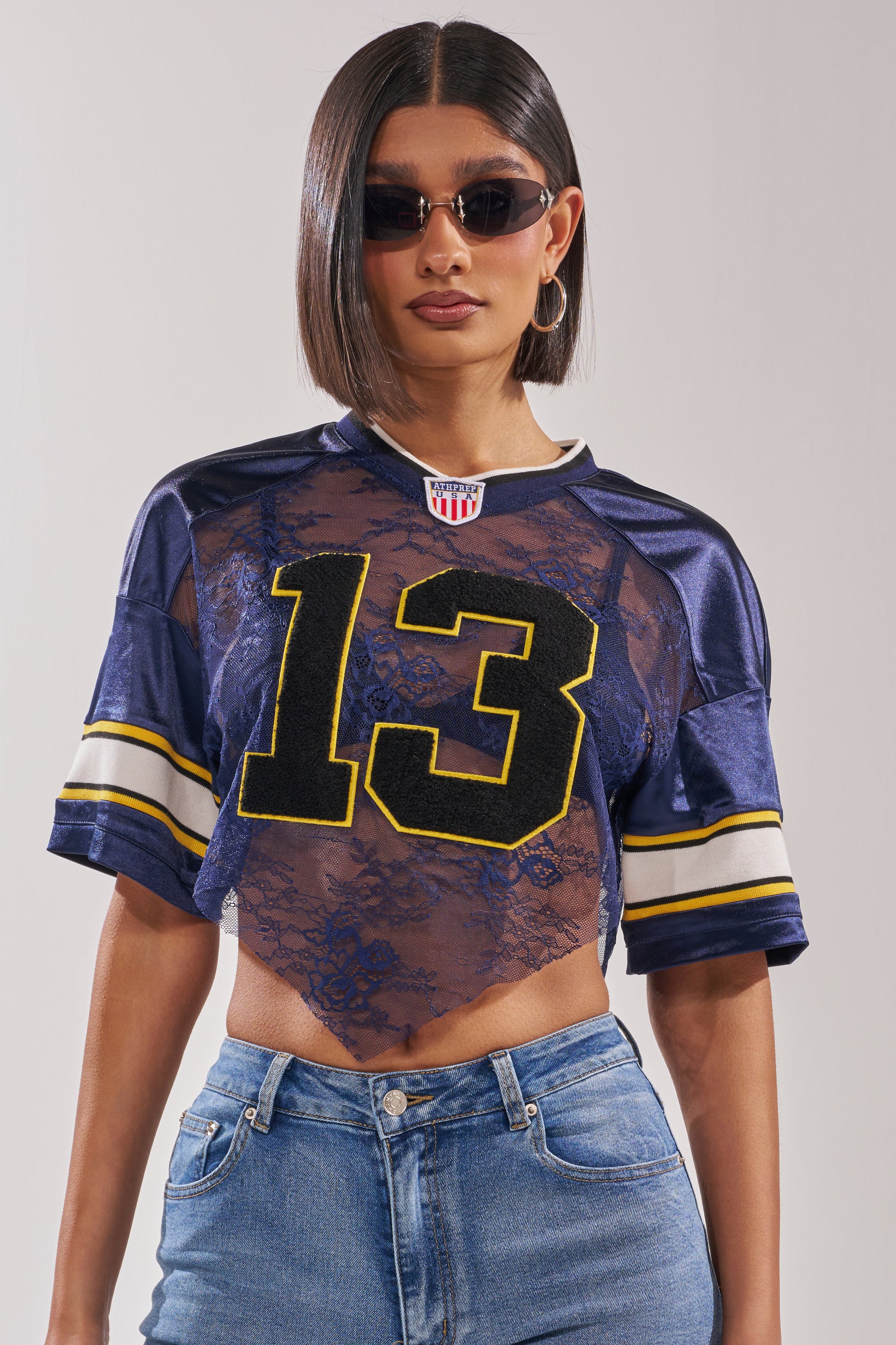 A woman in sunglasses and hoop earrings poses in the LUCKY LACE JERSEY TOP IN NAVY—featuring sheer navy lace, yellow stripes, number 13—paired with blue jeans. 3