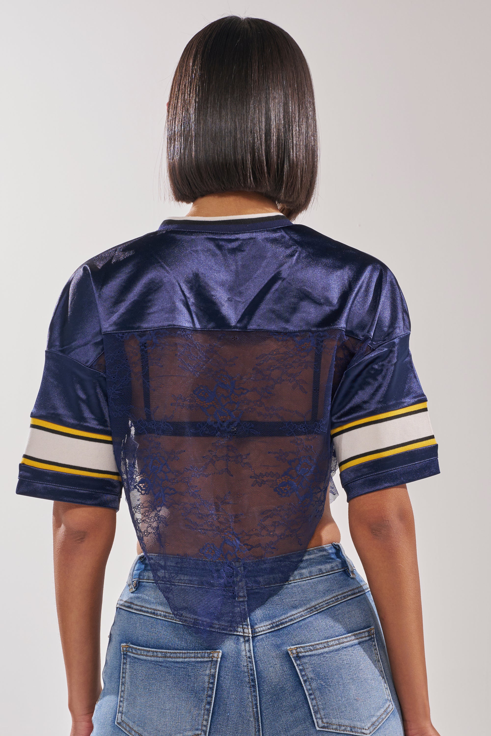 A person is seen from behind wearing the LUCKY LACE JERSEY TOP IN NAVY with striped sleeves and blue jeans. Their straight, shoulder-length hair partially covers the sporty streetwear top. 5