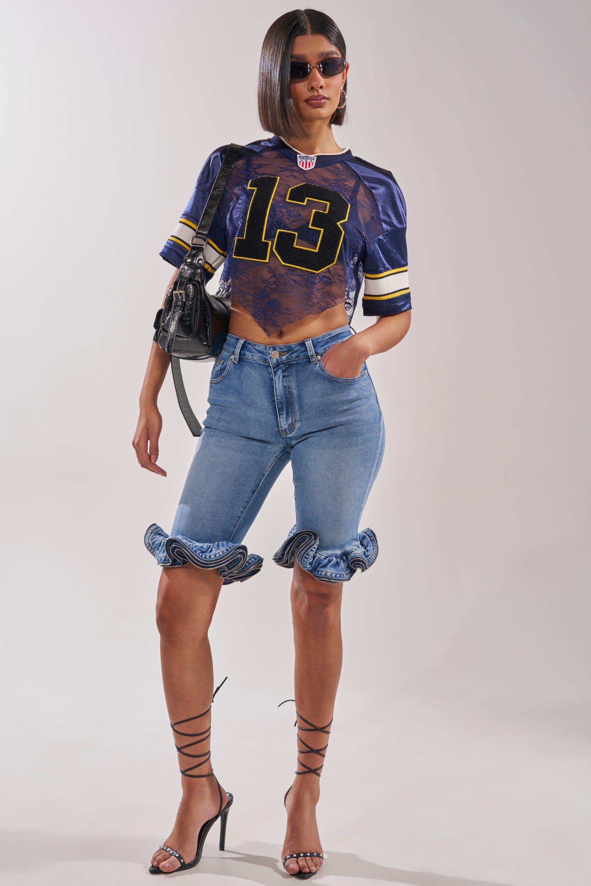 A woman poses confidently in the LUCKY LACE JERSEY TOP IN NAVY, paired with ruffled knee-length denim shorts, strappy high heels, sunglasses, and a black handbag. 6