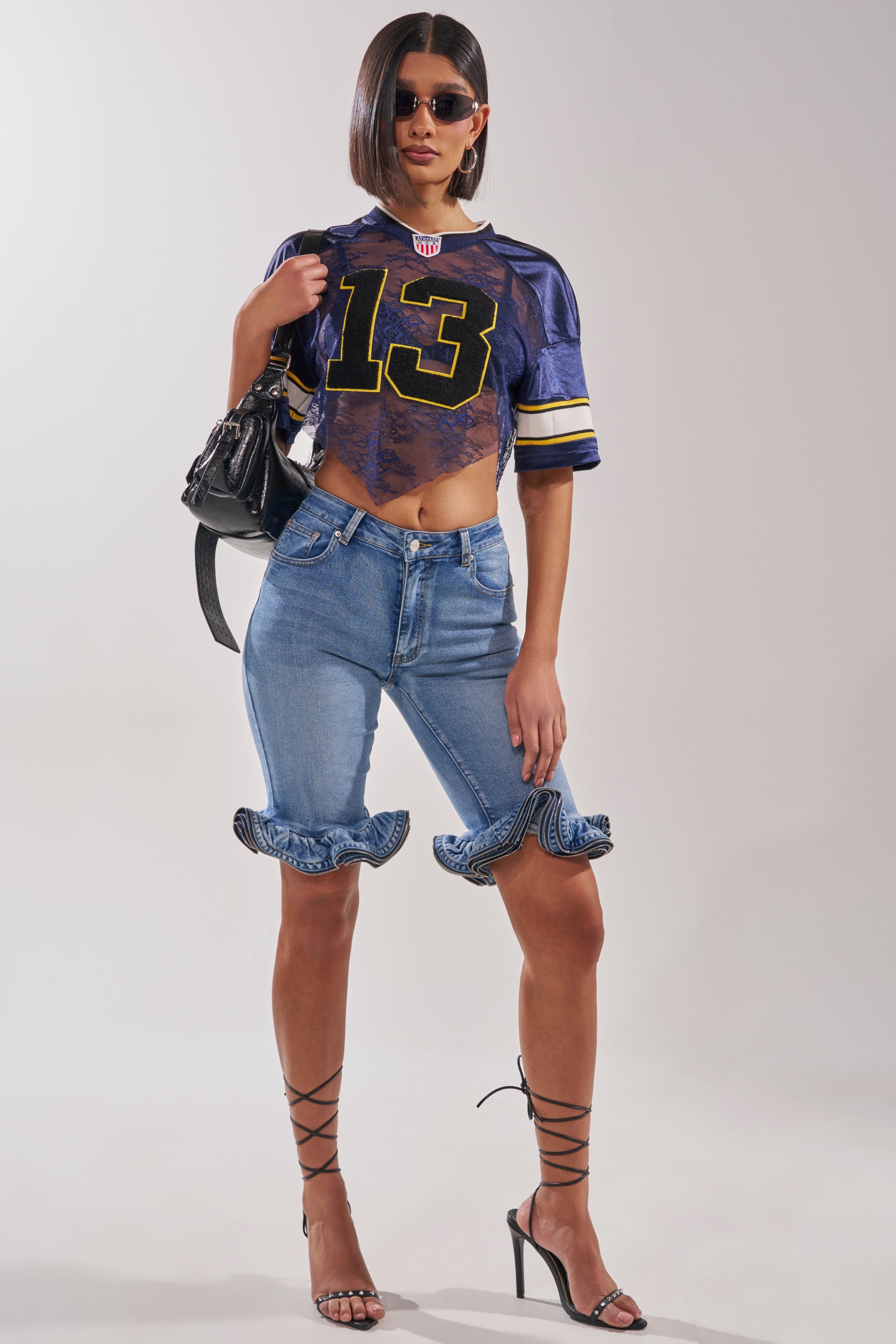 A woman poses confidently in sunglasses, the LUCKY LACE JERSEY TOP IN NAVY with the number 13, blue ruffled-hem denim shorts, black lace-up heels, and a black handbag slung over her shoulder. 7