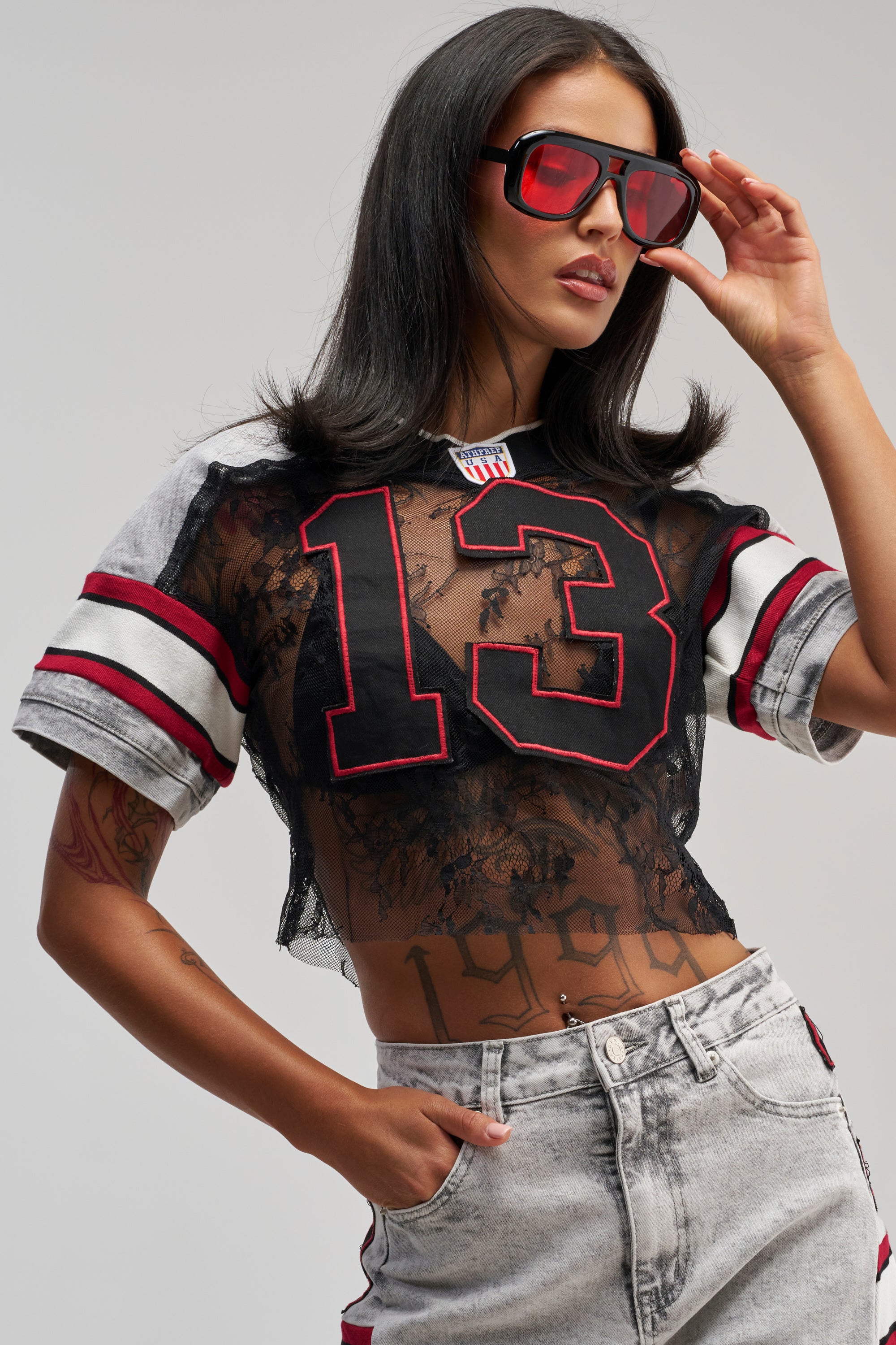 A woman with long dark hair and a confident pose wears red sunglasses, light gray jeans, and the VARSITY SQUAD JERSEY—a black lace varsity crop top featuring the number 13—showing off a “1998” tattoo on her midriff against a light background.