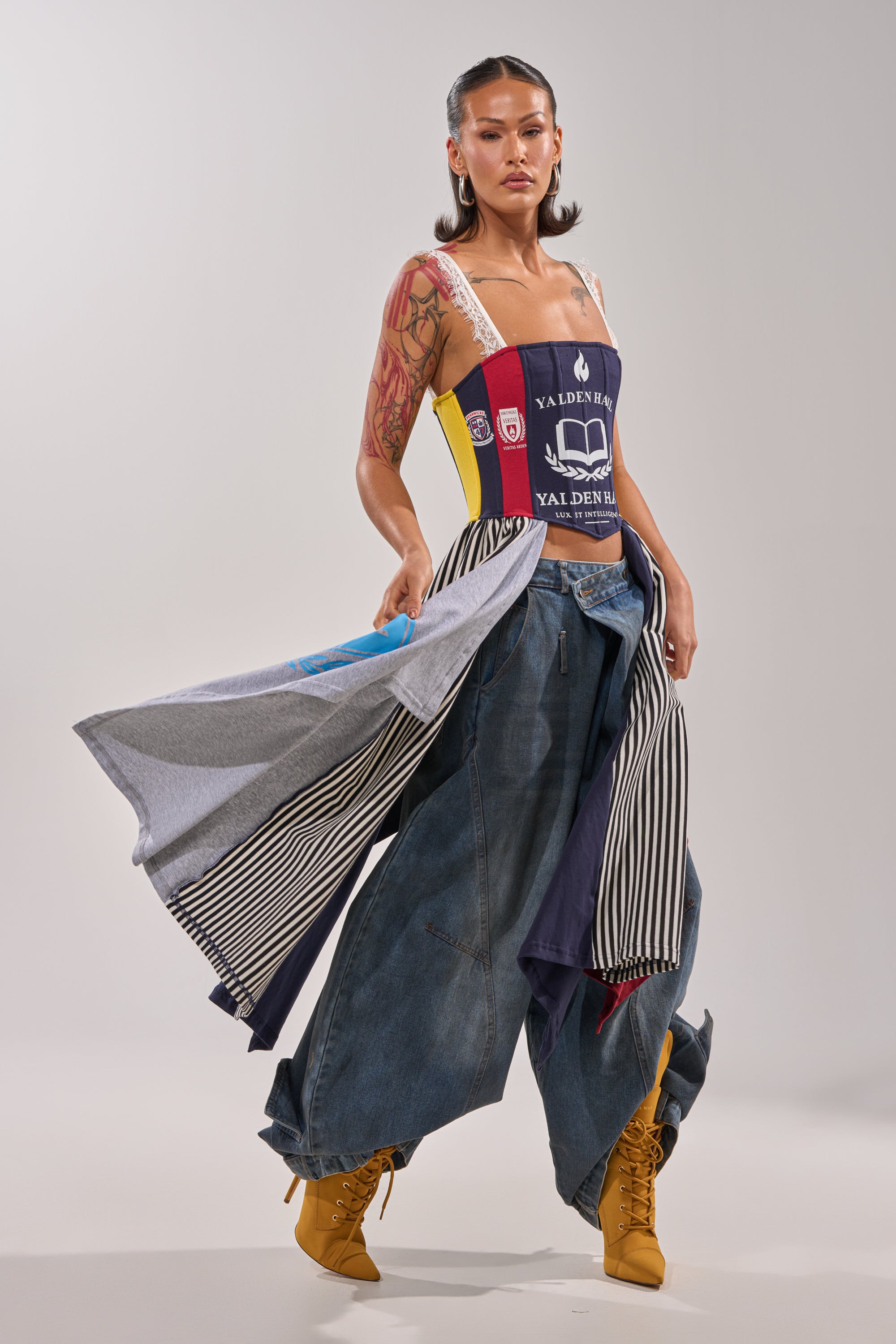 A woman exudes confidence in the UNIVERSAL CORSET paired with oversized baggy jeans, yellow lace-up boots, and a skirt of draped fabrics. Tattoos, hoop earrings, and slicked-back hair complete her bold festival look. 0