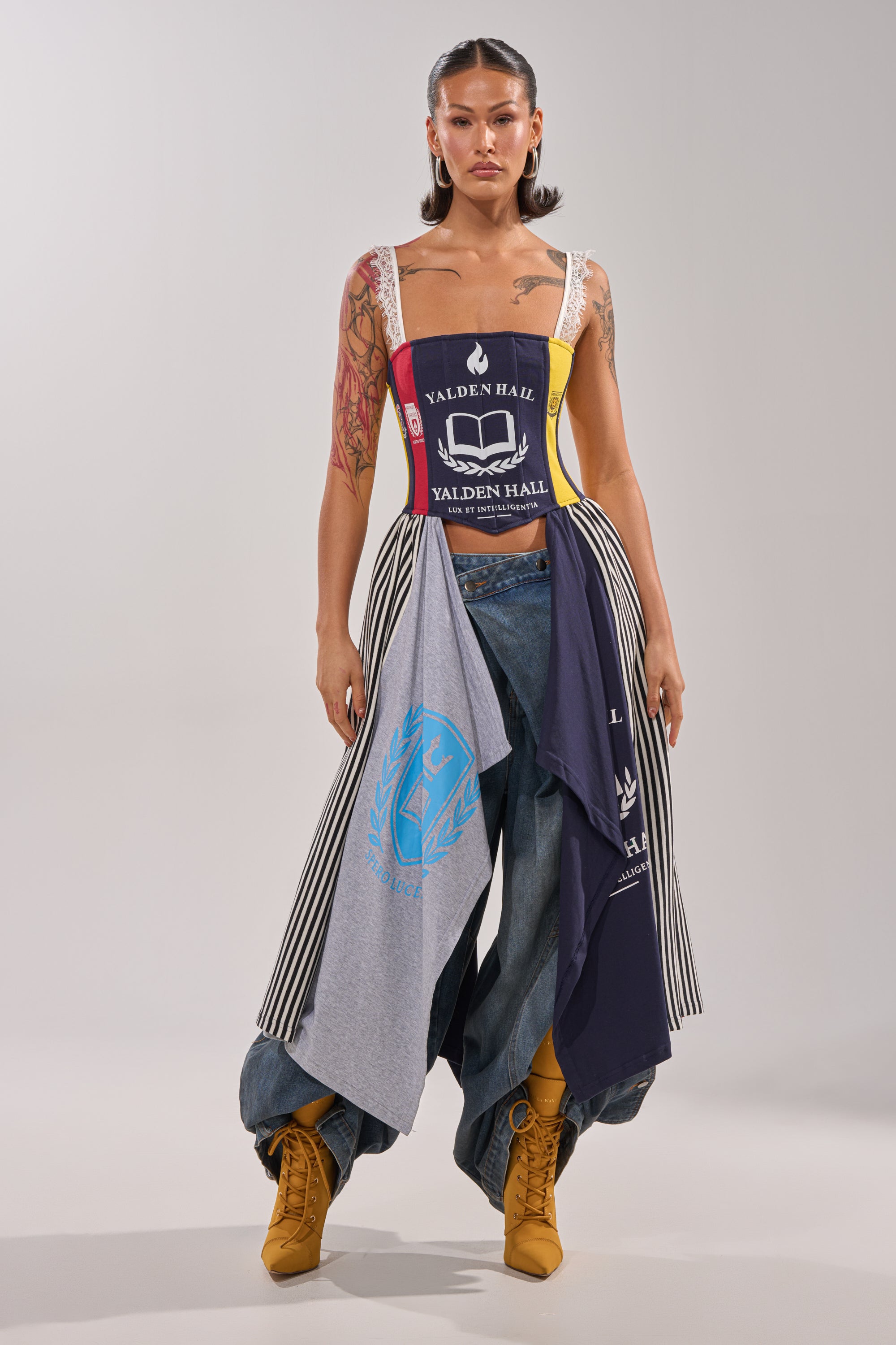 A woman stands confidently in festival attire, pairing the UNIVERSAL CORSET with wide-leg jeans, yellow boots, and a flowing skirt made from layered "Walden Hall" T-shirts, set against a plain light background. 1