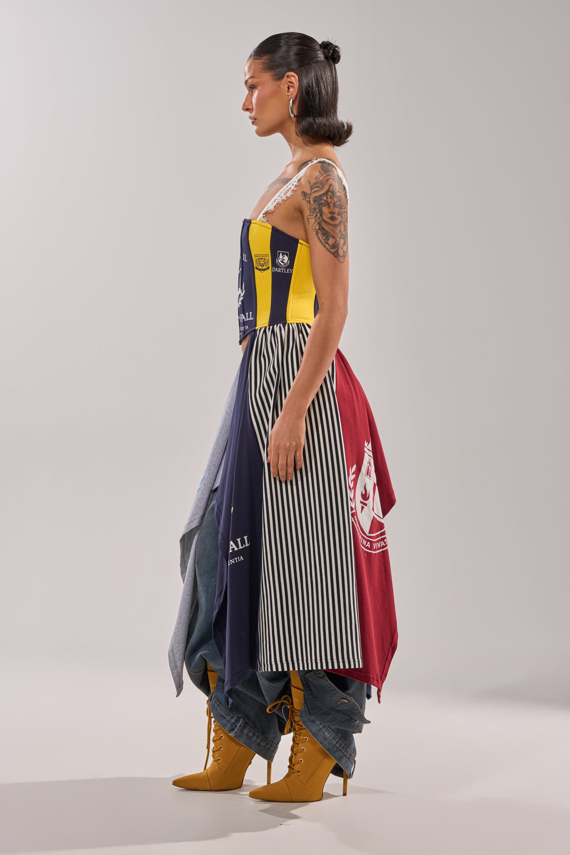 A person with shoulder-length dark hair stands in profile, wearing the UNIVERSAL CORSET—a colorful patchwork piece with stripes, logos, and patterns—styled with layered denim pants and yellow lace-up heeled boots for a bold streetwear look. 3