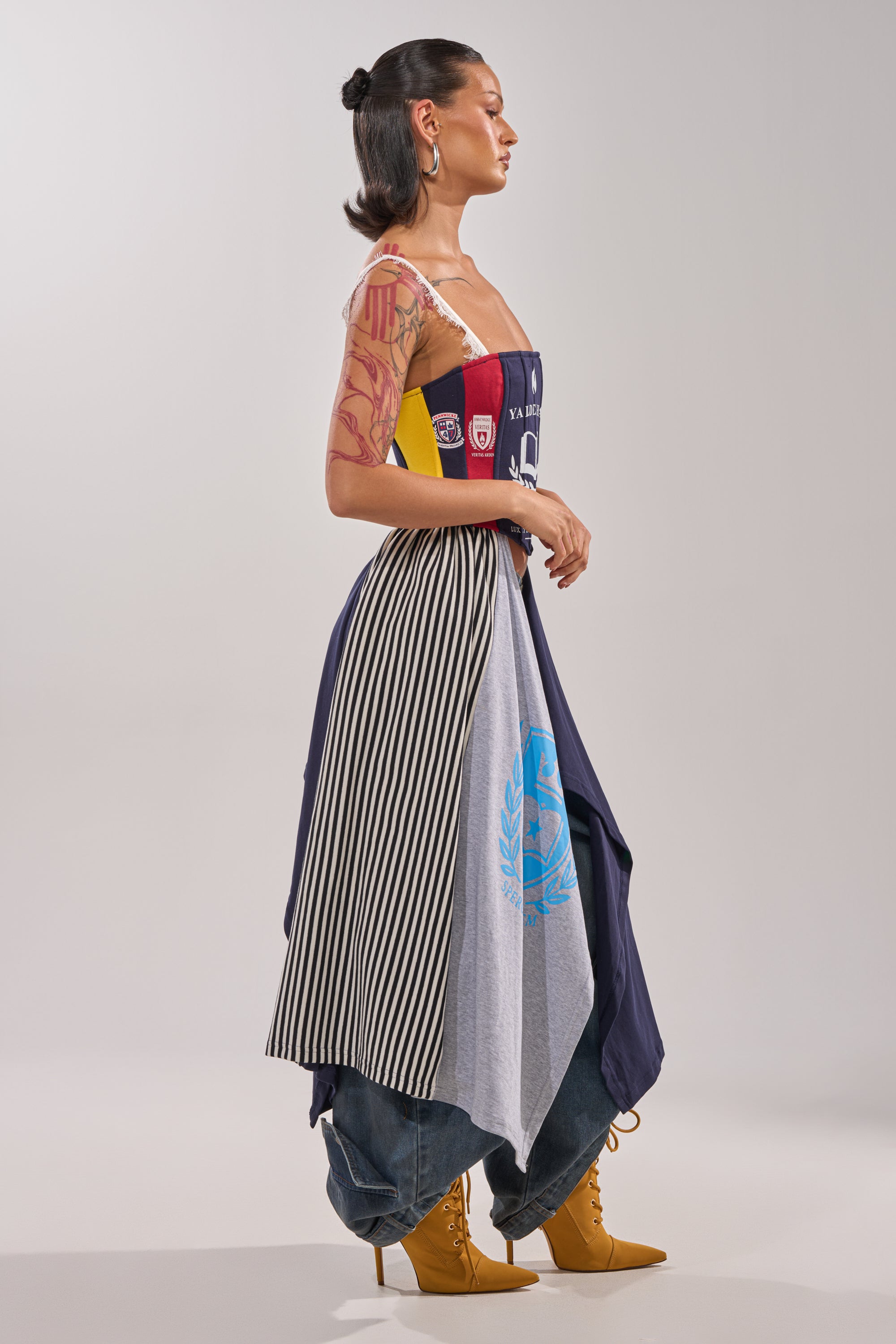 A woman in profile sports the UNIVERSAL CORSET over a multicolored patchwork dress and striped layers, styled with blue jeans and tan lace-up boots. She channels street style with visible arm tattoos and a sleek dark bun. 4