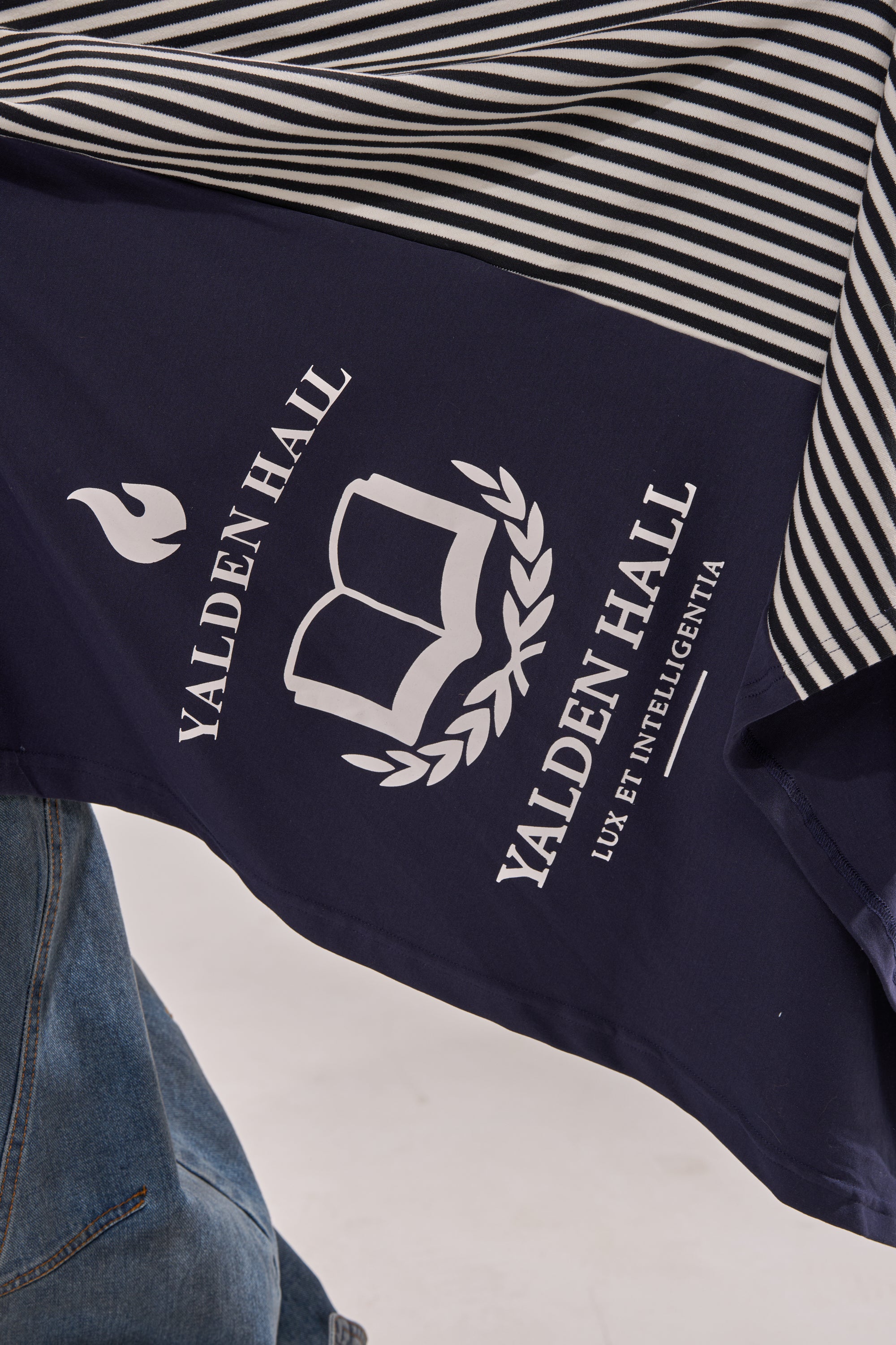 The UNIVERSAL CORSET is a navy blue and white striped streetwear piece with a crest of an open book, flame, laurel branches, and the text "VALDEN HALL" and "LUX ET INTELLIGENTIA," shown next to blue jeans. 6