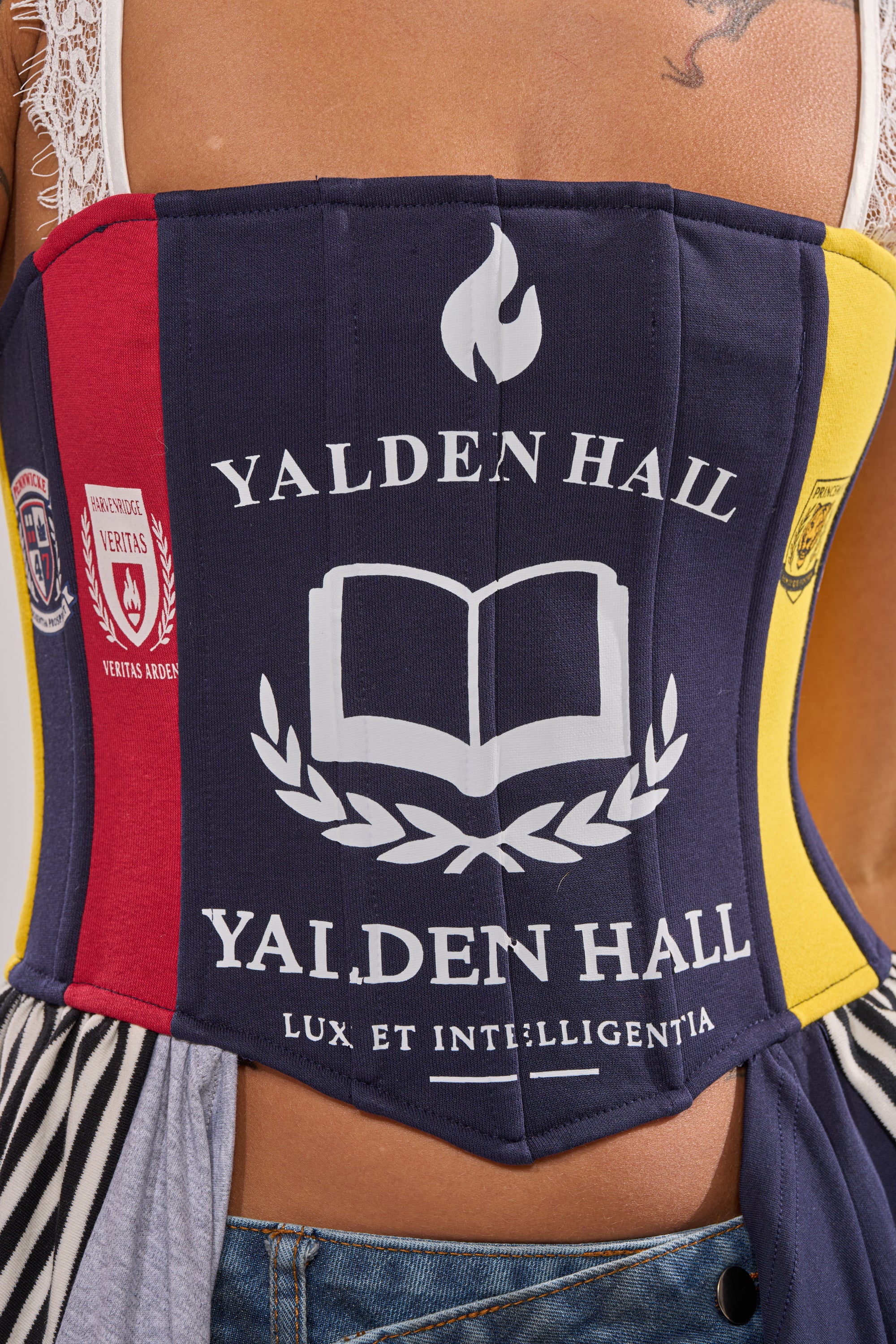 A close-up of someone in the UNIVERSAL CORSET—a colorful statement top with patches, "Yalden Hall" above an open book emblem, and "Lux et Intelligentia" motto. White lace straps and a tattoo peek from their upper back, ideal for festival styling. 7
