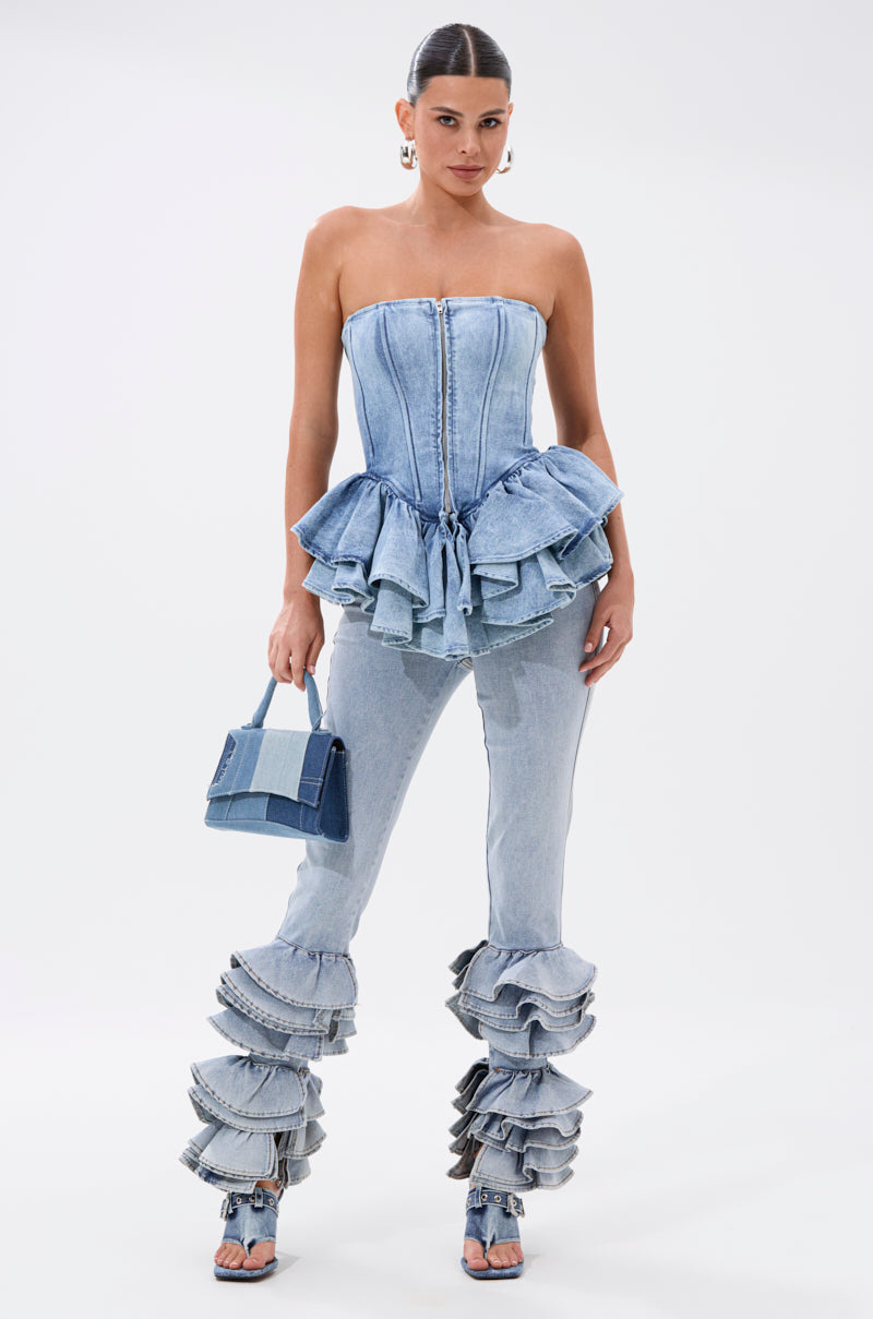 A woman in the EVA DENIM PEPLUM CORSET TOP with ruffled hem pairs it with matching denim pants, open-toe heels, and a small blue handbag, posing confidently against a plain white background.