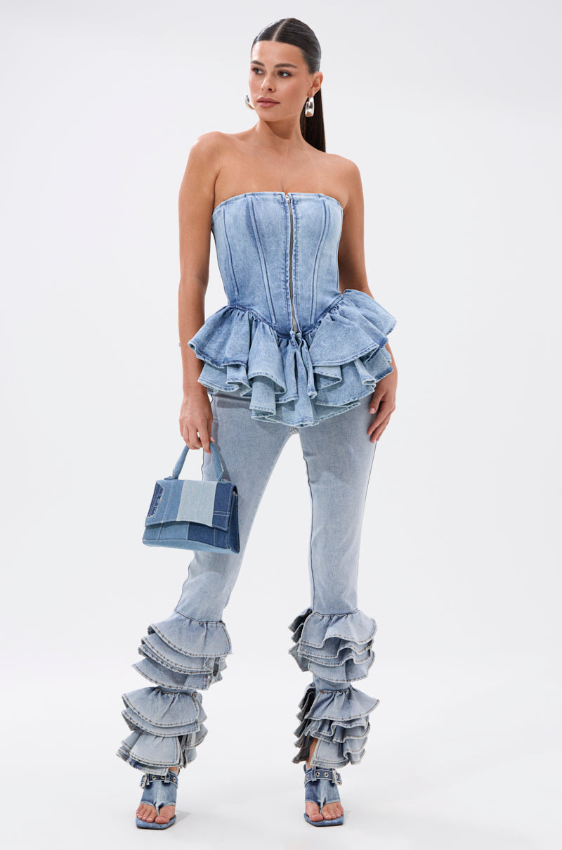 A woman wears the EVA DENIM PEPLUM CORSET TOP with a ruffled peplum hem, matching layered-ruffle denim pants, denim sandals, and holds a small denim handbag against a white background.
