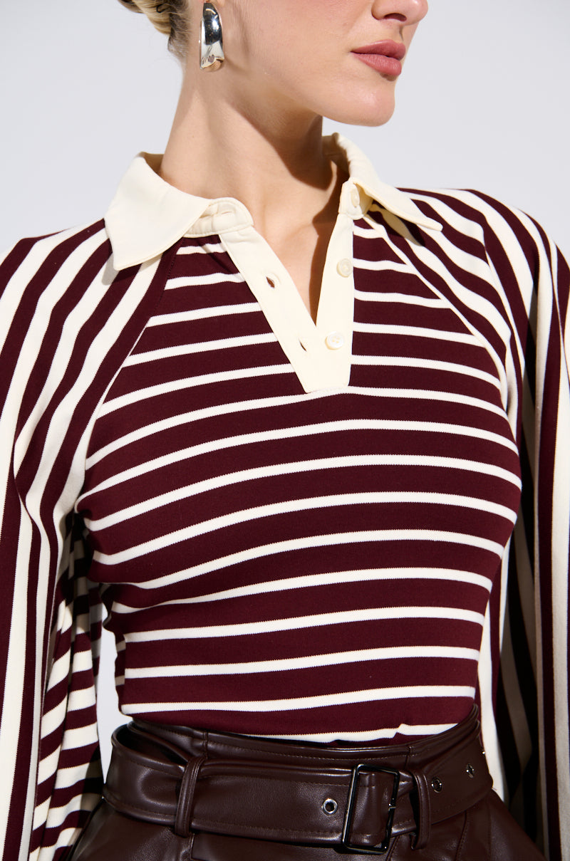 A woman wears the ALMA STRIPE COLLARED BODYSUIT in burgundy and cream with cream buttons, paired with brown belted high-waisted pants and silver geometric earrings. Looking to the side, she highlights the bold stripes of the bodysuit.